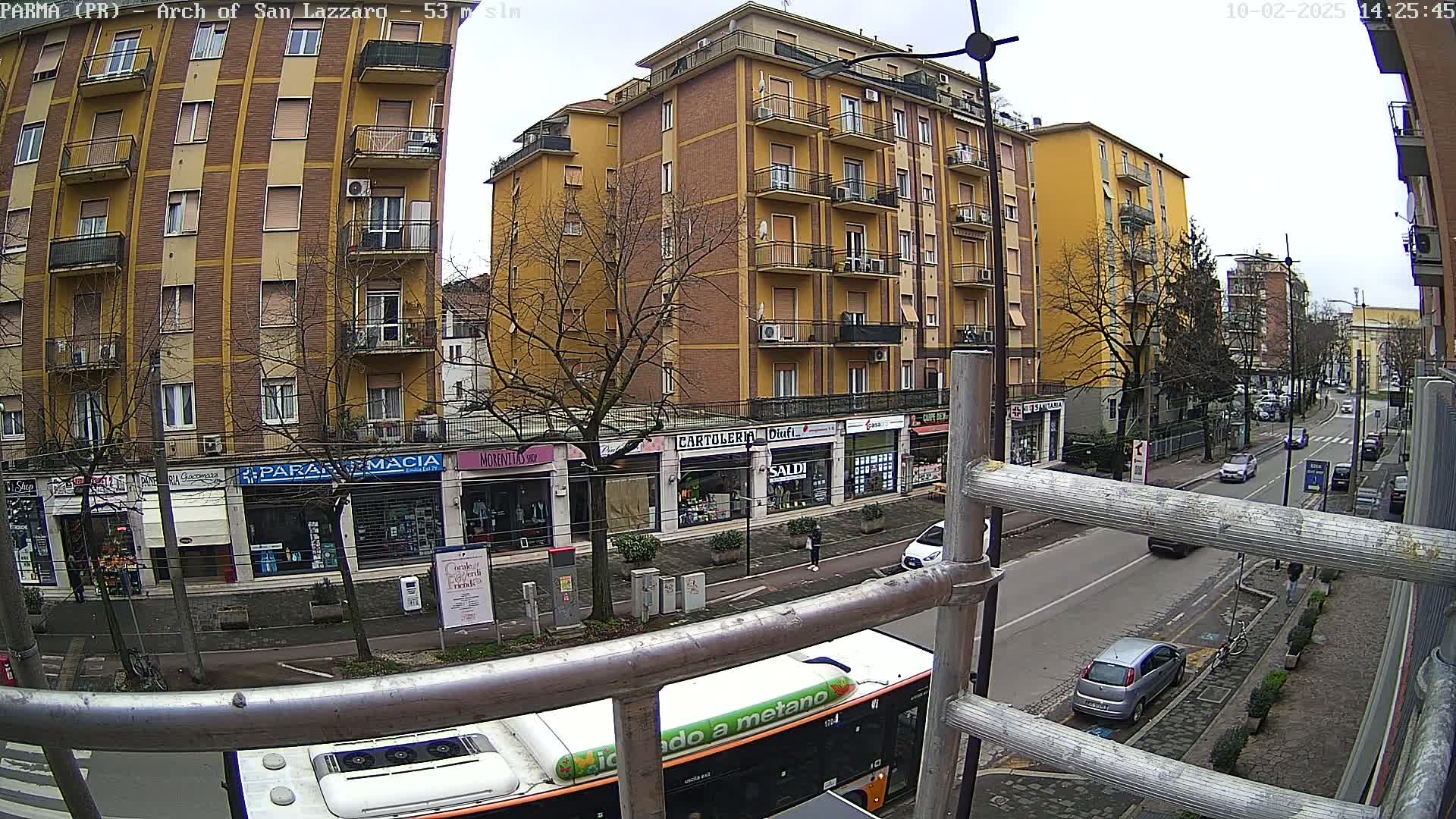 A city street scene on an overcast day shows a multi-story yellow building with shops on the ground floor, several cars on the street, and a portion of a bus visible behind scaffolding.