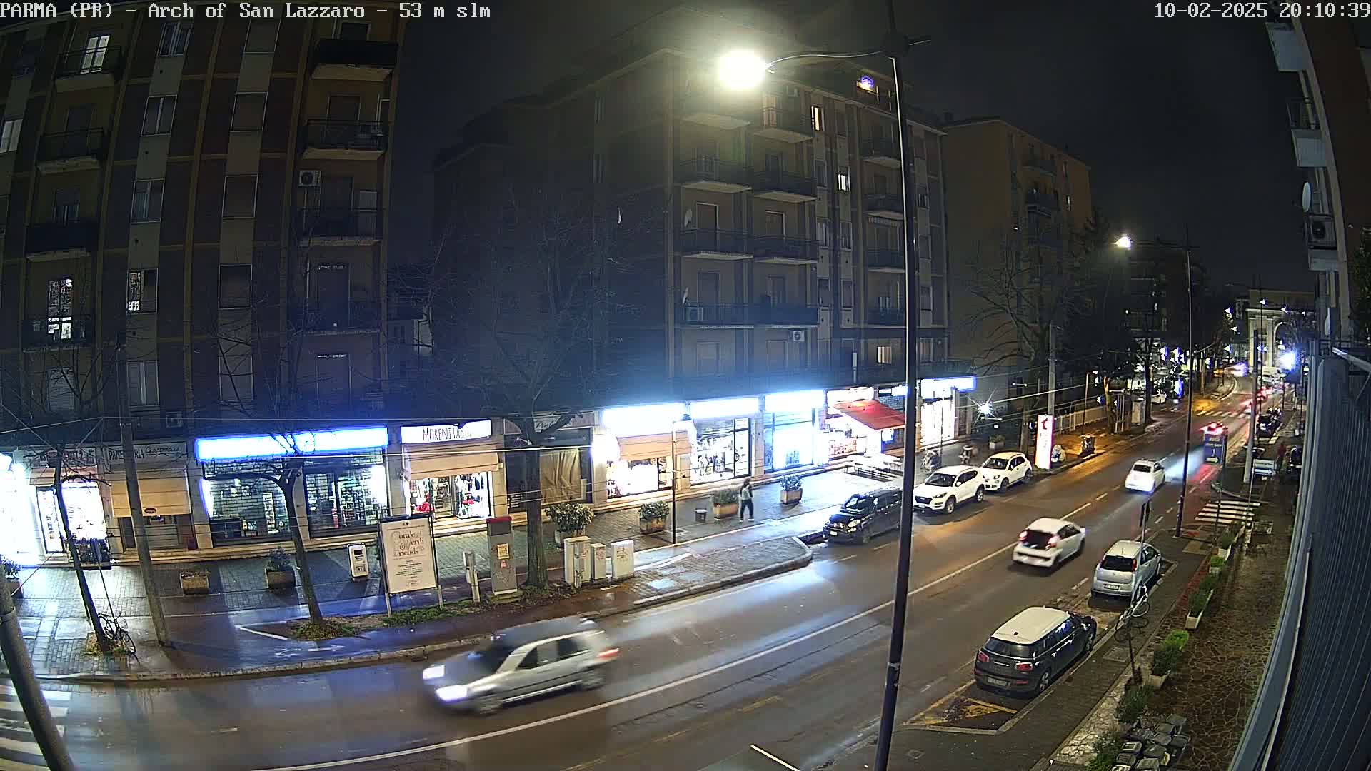A wet, nighttime street scene shows several cars driving past closed shops and a multi-story building.