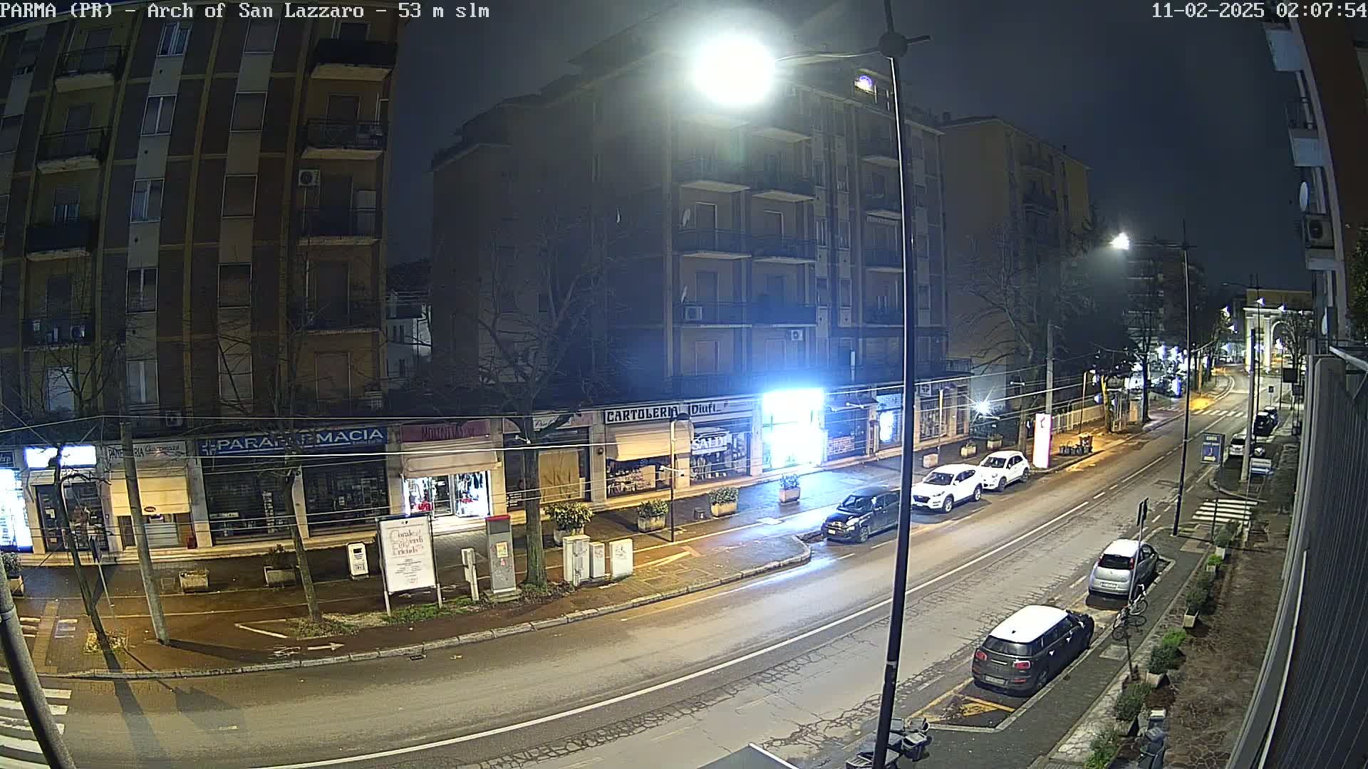 A nighttime street scene shows a row of shops and a multi-story building under streetlights, with several cars parked along the wet road.
