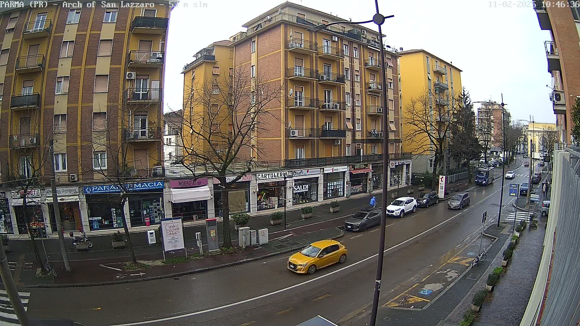 A wet city street lined with yellow buildings and shops, with cars parked along the side and a few cars driving down the street on an overcast day.