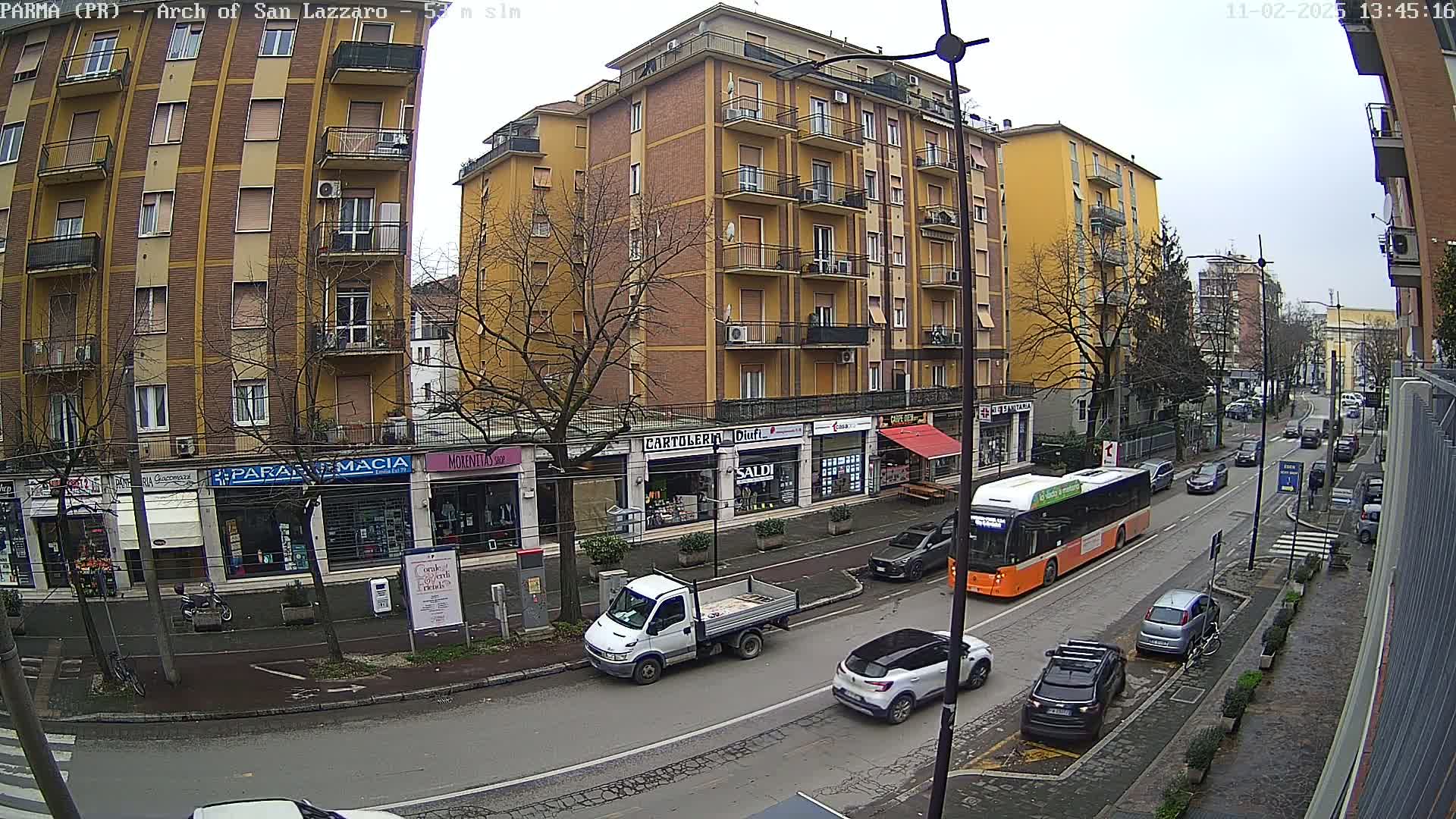 A street scene on an overcast day shows a multi-story building with shops on the ground floor, several cars and a bus on the road, and bare trees lining the street.
