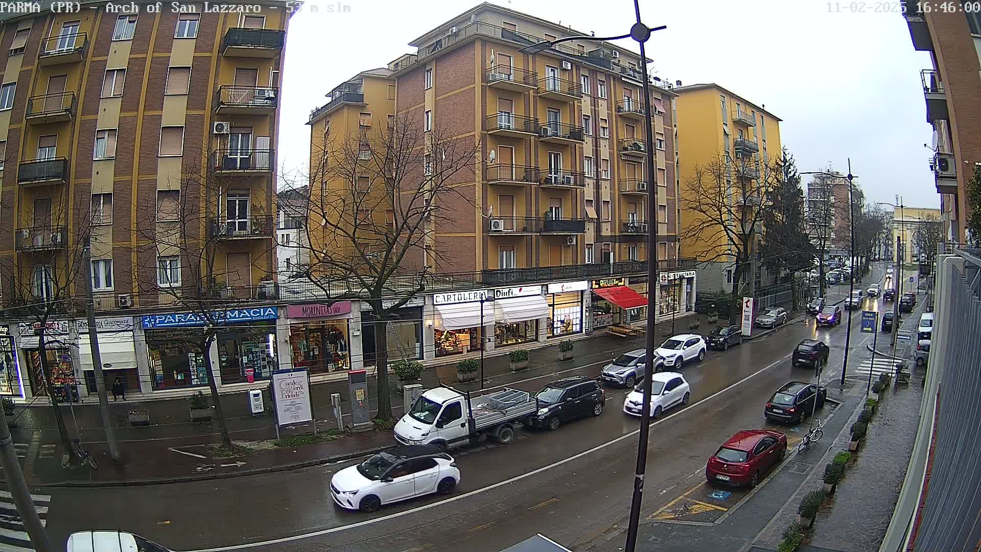 A wet city street scene shows a multi-story building with shops on the ground floor, and several cars parked and driving on a rainy day.