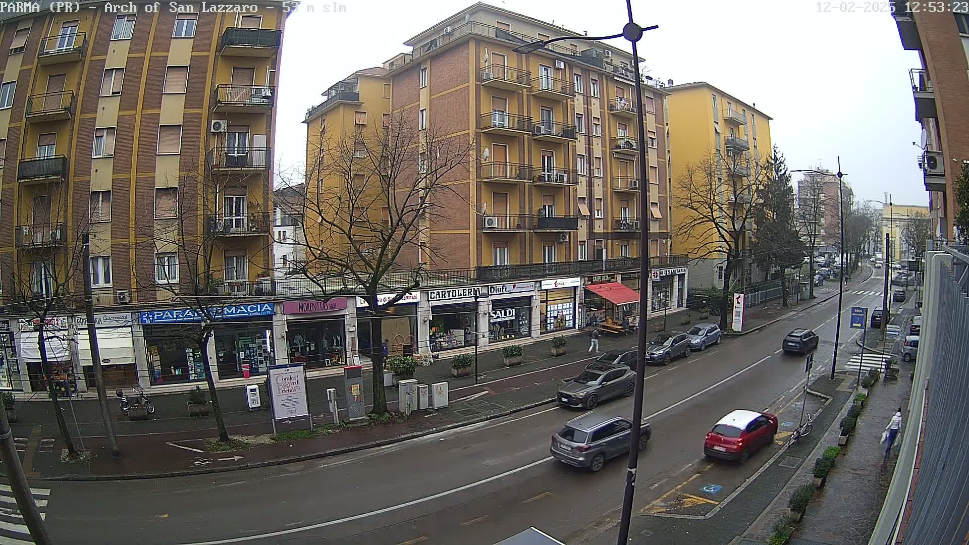 A street scene on an overcast day shows a multi-story yellow building with shops on the ground floor, several cars parked and driving on the street, and bare trees along the sidewalk.