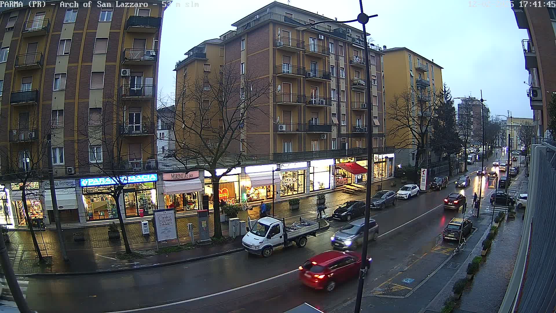 A wet street scene shows several cars driving past shops and a multi-story building under an overcast sky.