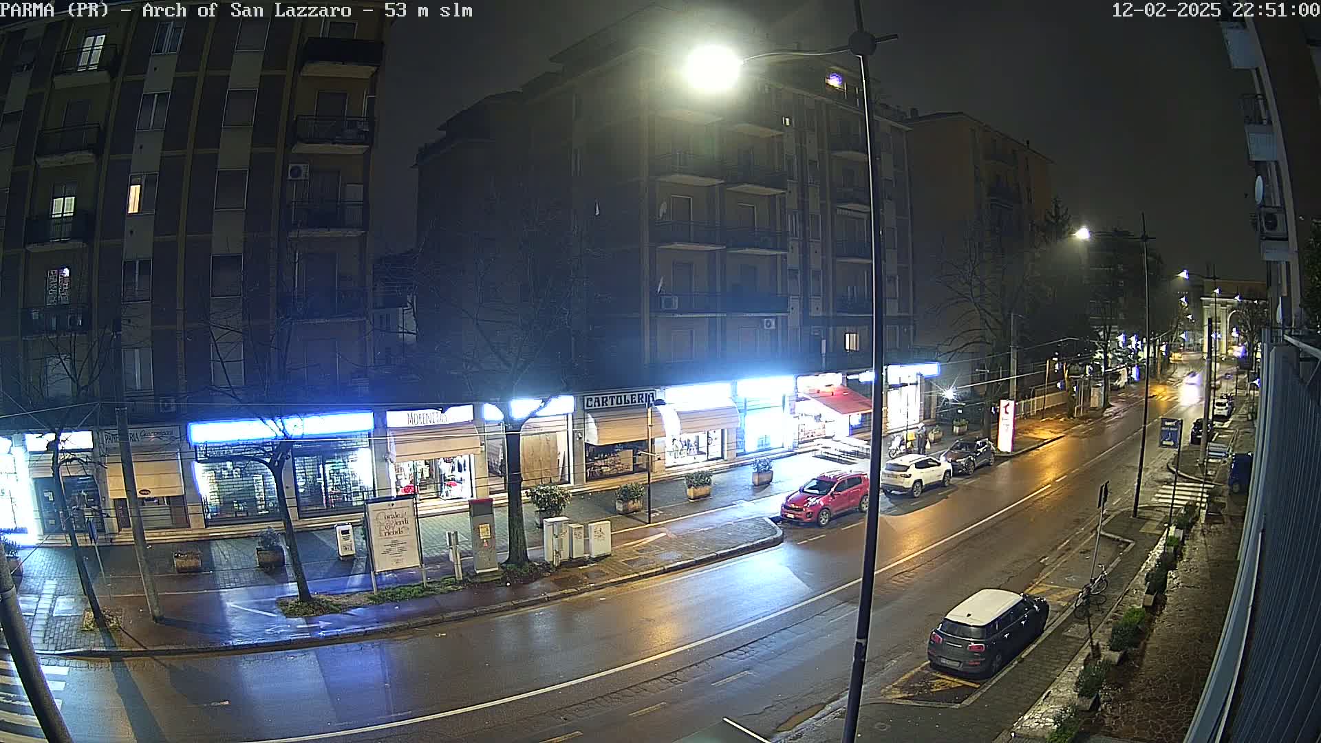 A wet, nighttime street scene shows several cars parked and driving past shops with illuminated signs, under bright streetlights.