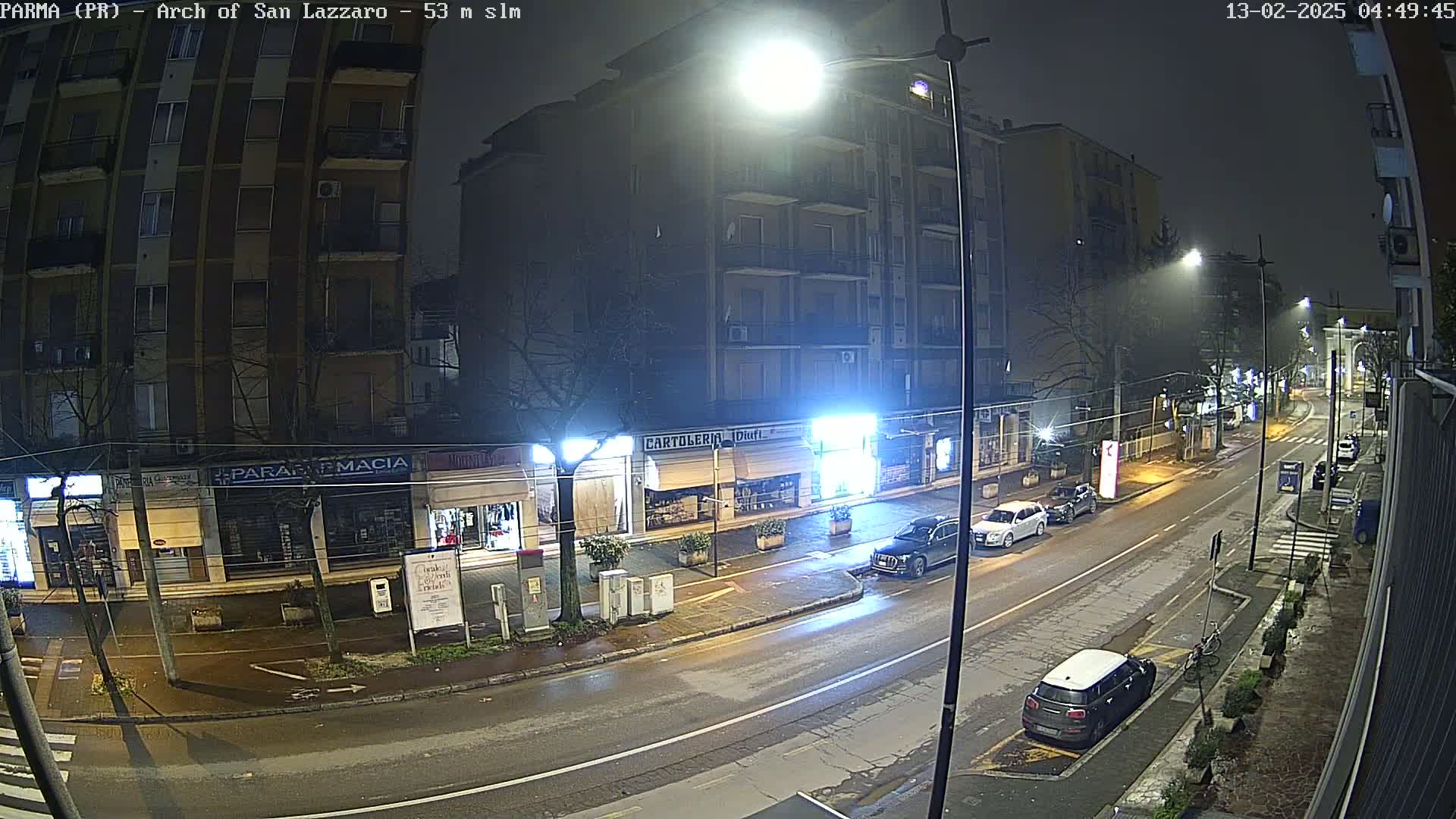 A nighttime street scene shows several parked cars and a few driving cars on a wet road, with shops and a multi-story apartment building lining the street under bright streetlights in foggy conditions.