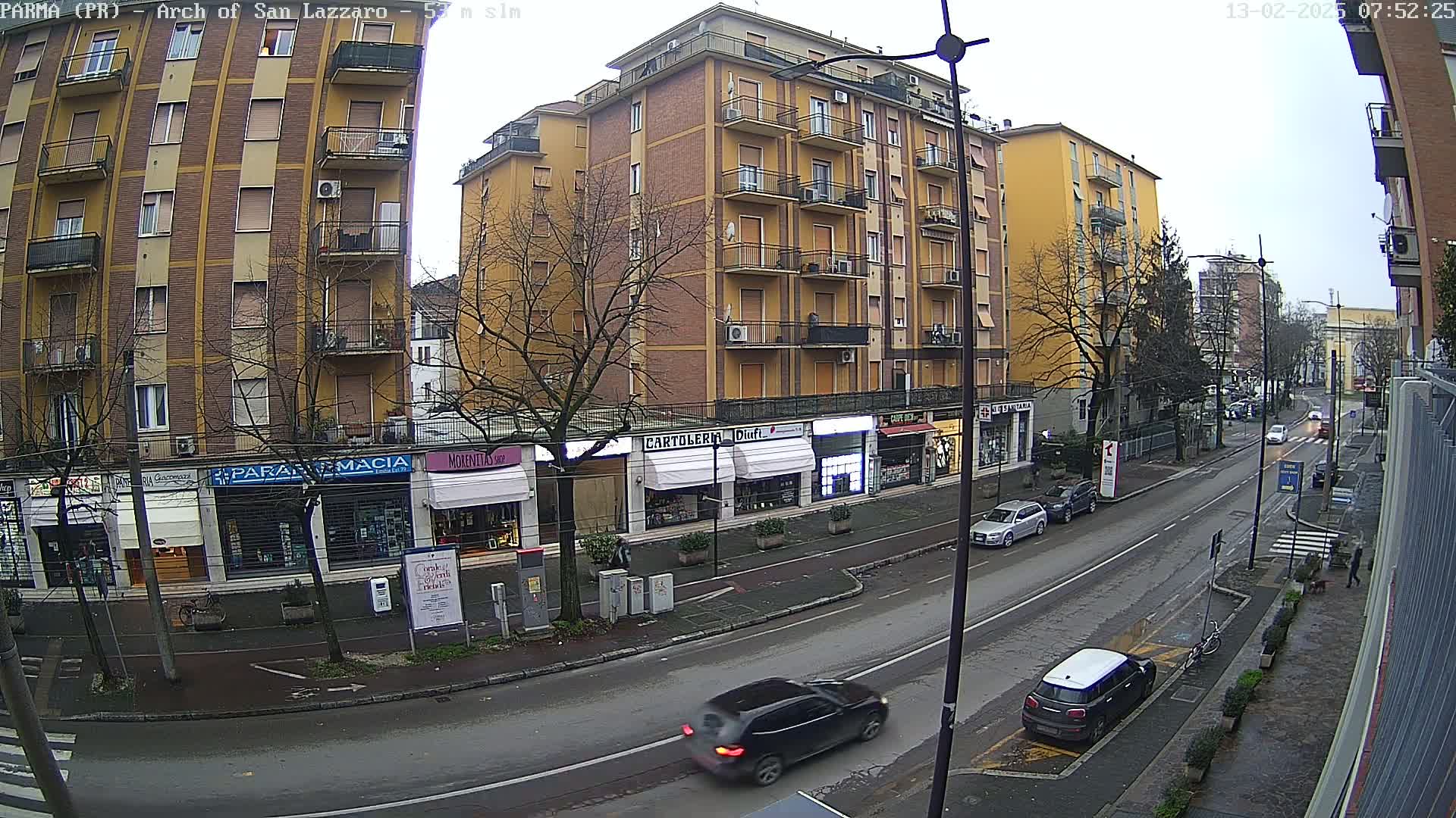 A street scene on an overcast day shows several multi-story buildings with shops on the ground floor, and several cars parked and driving down the street.