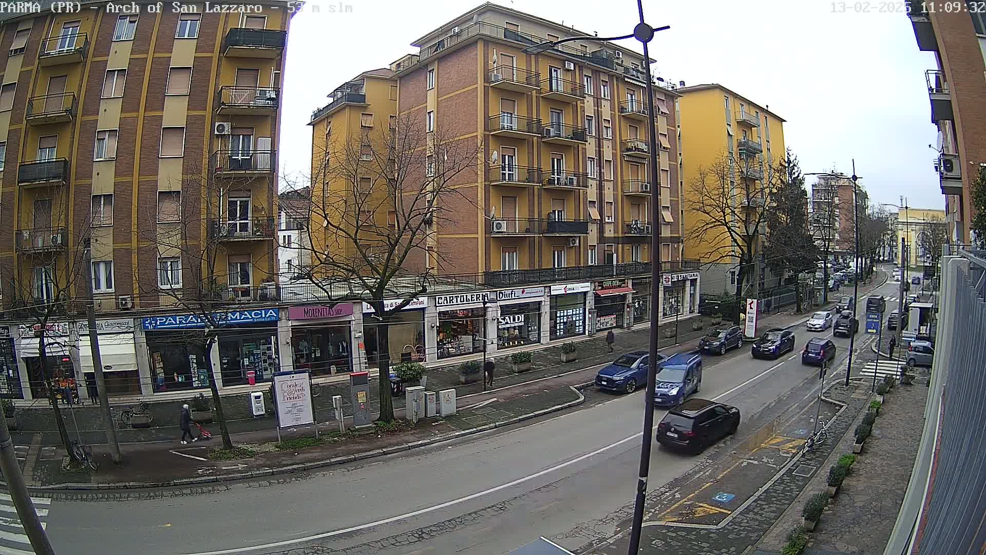 A street scene on an overcast day shows several multi-story buildings with shops on the ground floor, and several cars driving down a street.