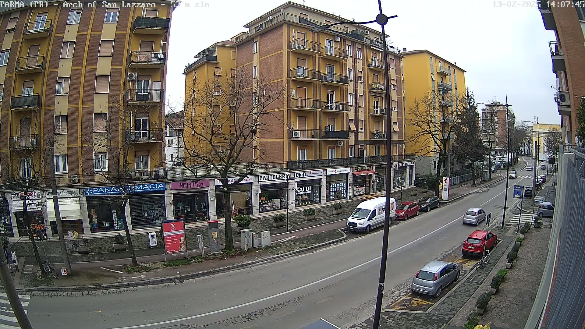 A street scene on an overcast day shows a multi-story yellow building with shops on the ground floor, several parked cars, and a few cars driving down the street.