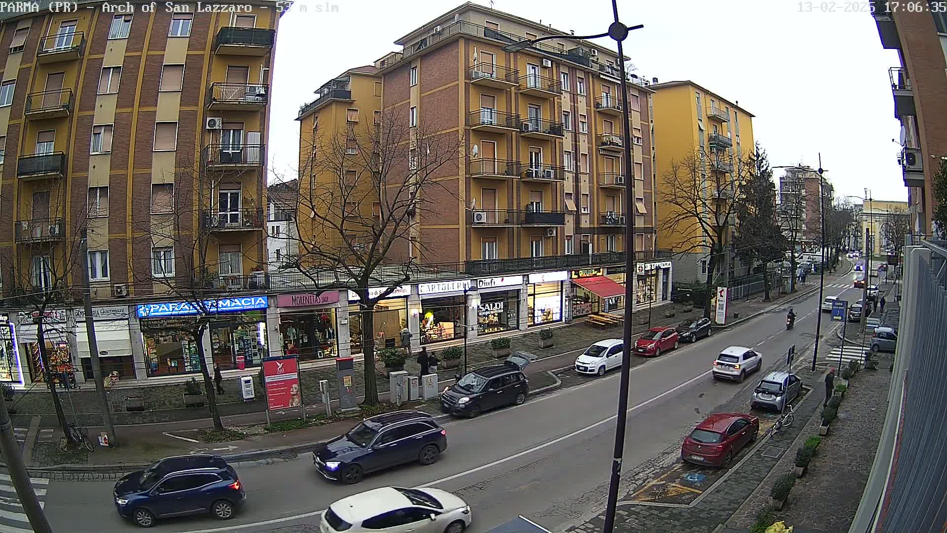 A street scene on an overcast day shows a row of shops and multi-story buildings, with cars parked and driving along a road.