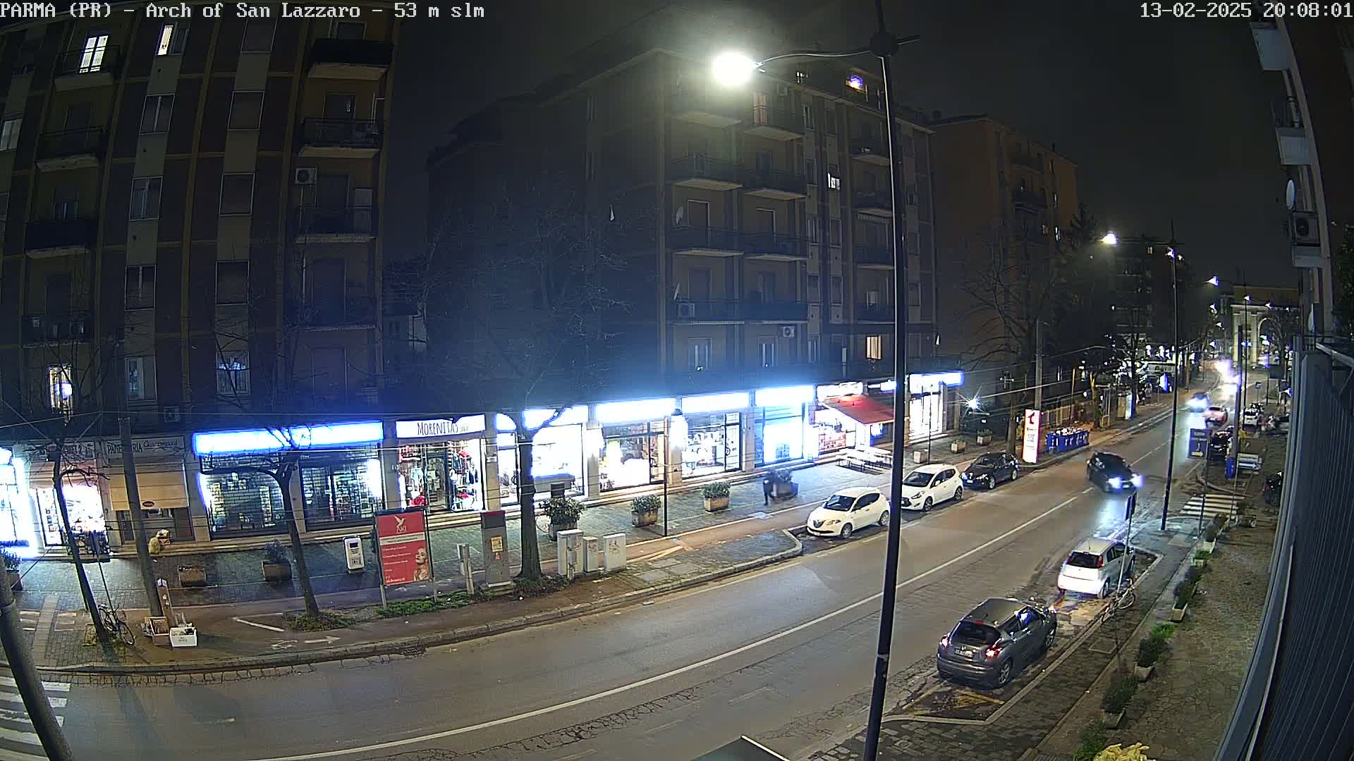 A nighttime street scene shows several cars parked and driving along a roadway lined with storefronts and apartment buildings under streetlights.
