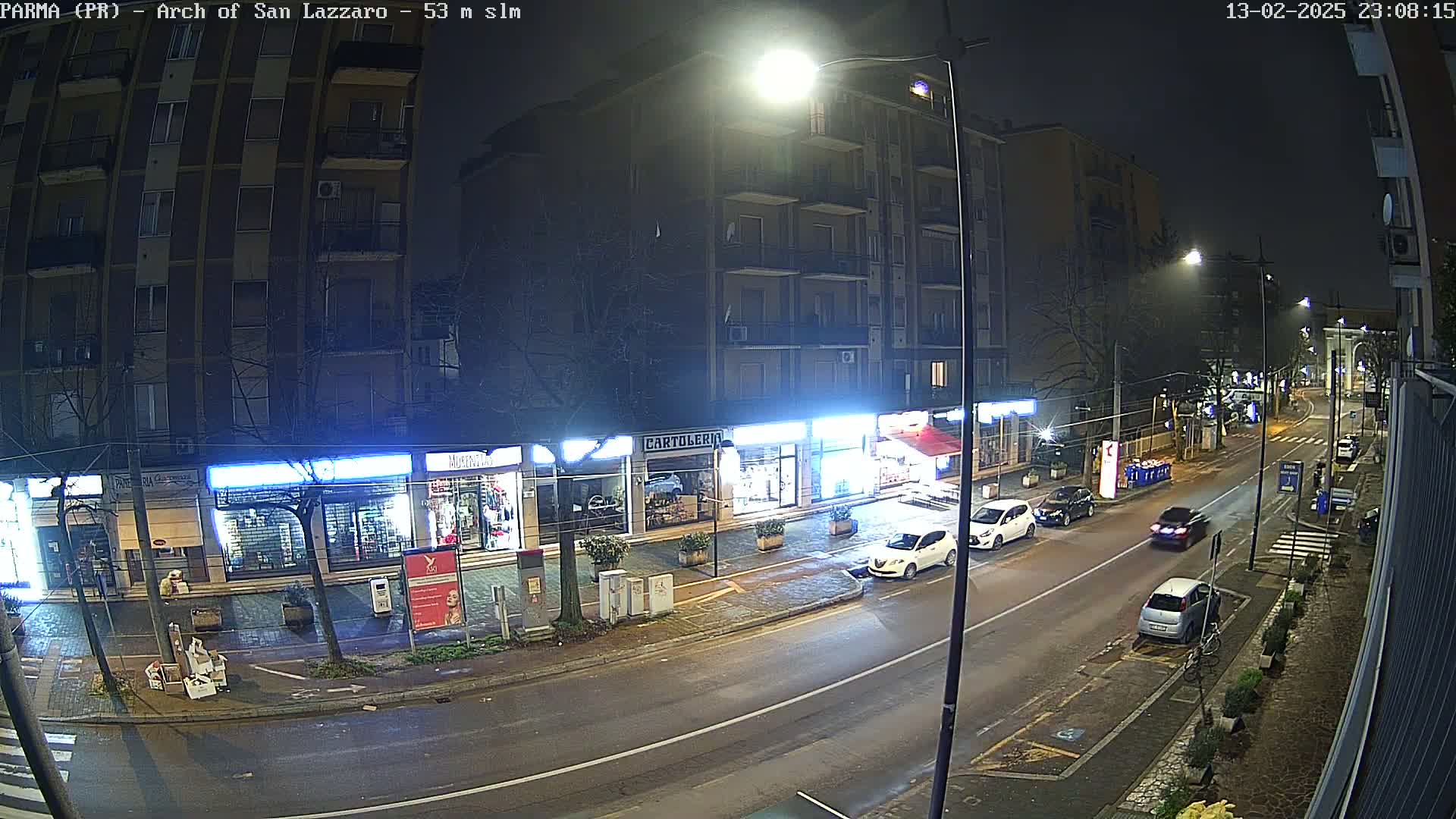 A nighttime street scene shows several cars parked and driving along a wet road in front of various shops and a multi-story building.