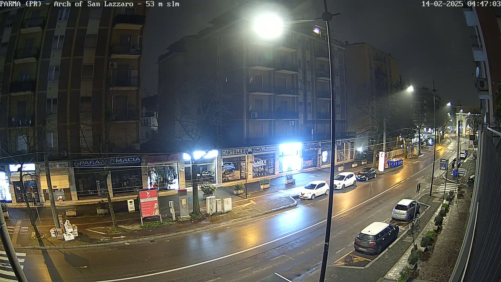 A wet, nighttime street scene shows several parked cars and a few driving cars near a row of shops under bright streetlights.