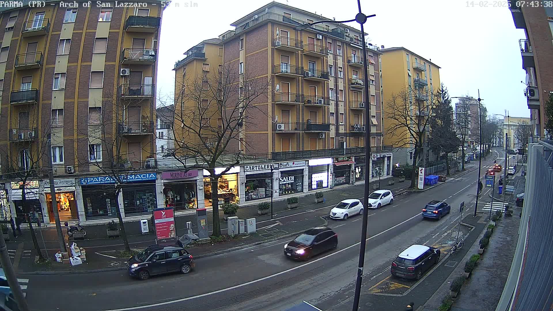 A street scene on an overcast day shows several cars driving past multi-story buildings with ground-floor shops.