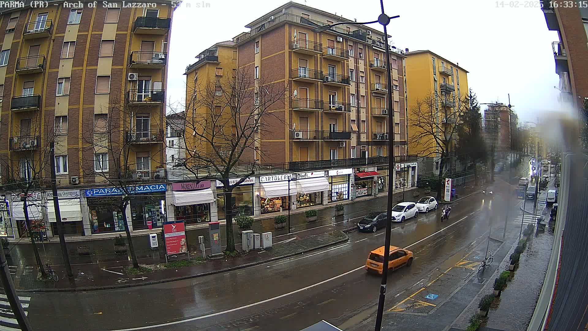 A rain-slicked street scene shows several cars parked and driving past multi-story buildings with shops on the ground floor under a gray, overcast sky.
