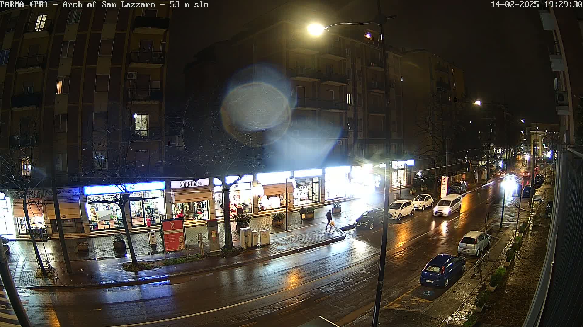 A rain-slicked city street at night, showing several parked and moving cars in front of illuminated storefronts and a multi-story apartment building.