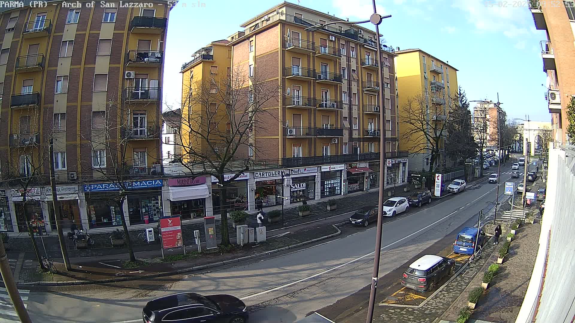 A sunny day on a city street lined with multi-story buildings and shops, with cars parked along the curb and a few cars driving on the road.