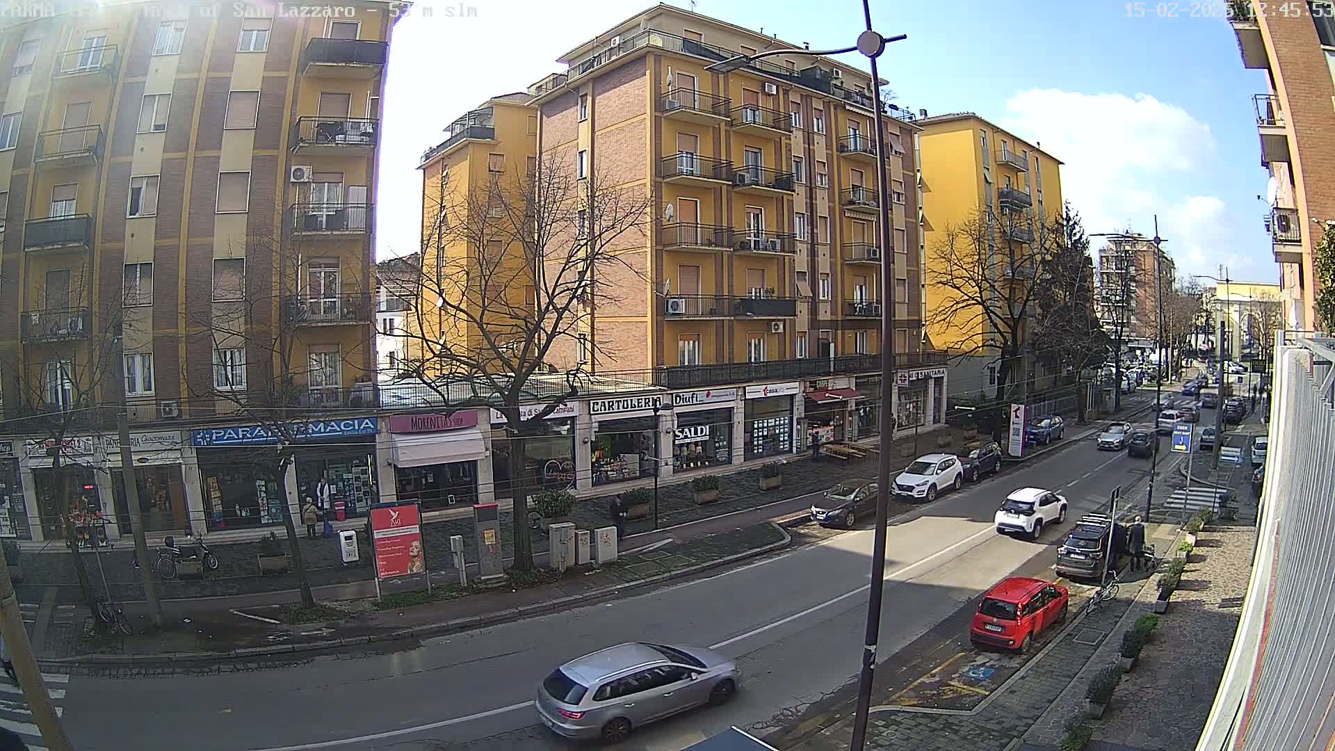 A sunny street scene shows a multi-story yellow building with shops on the ground floor, along with parked cars and cars driving on the road.