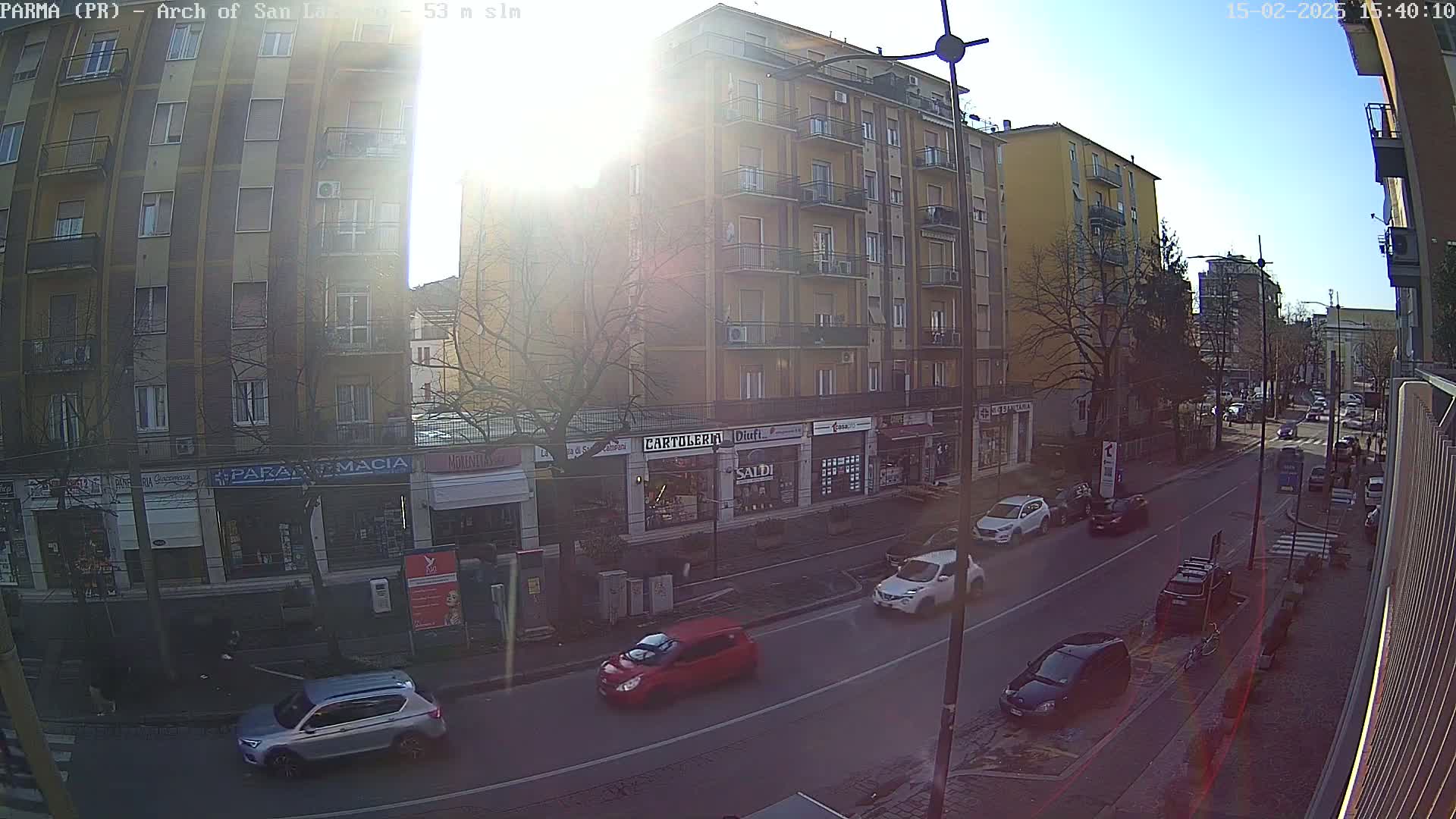 A sunny day view down a city street lined with multi-story buildings and shops, with several cars parked and driving along the road.