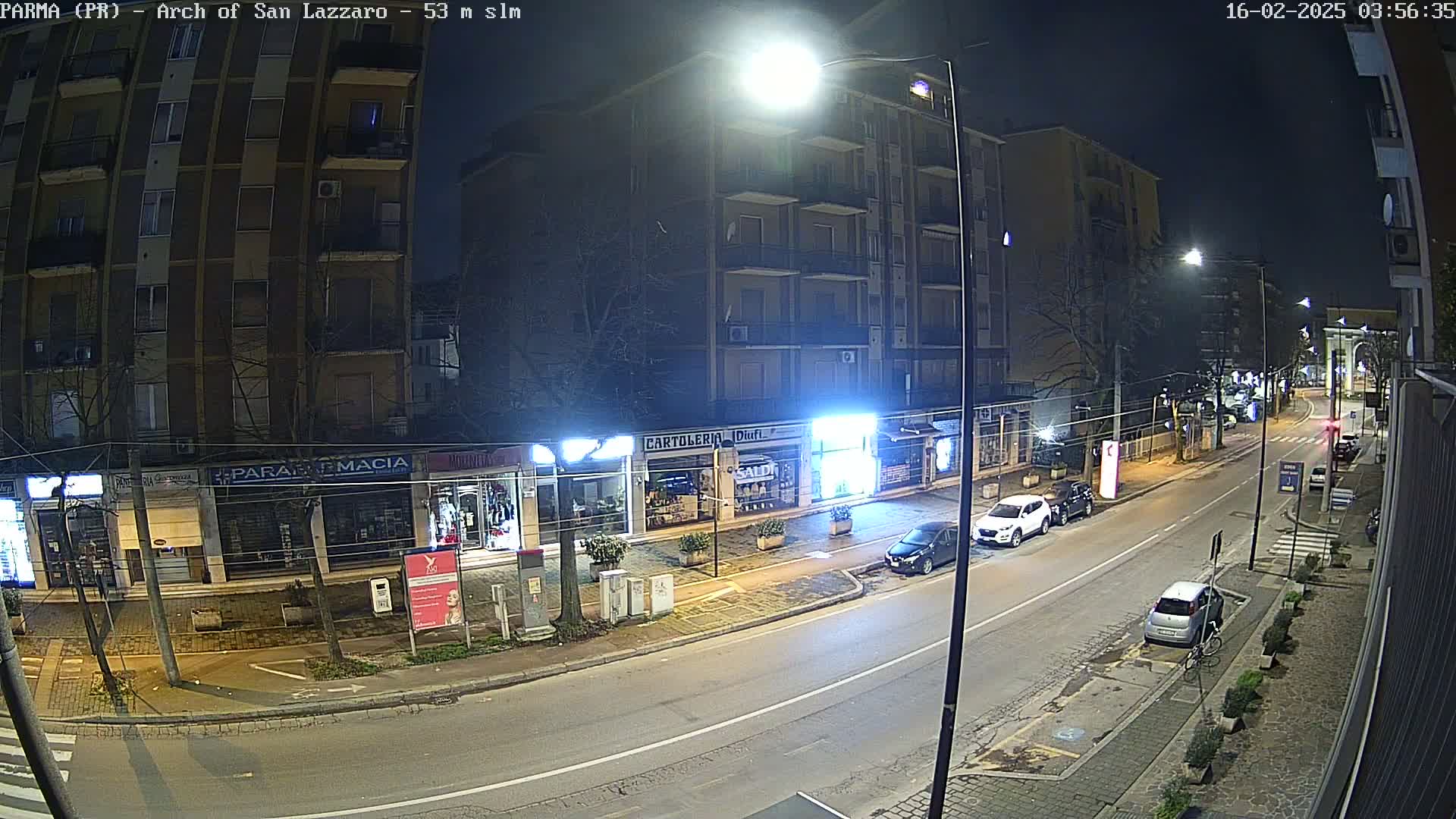 A nighttime street scene shows a sparsely populated road lined with shops and apartment buildings under bright streetlights.