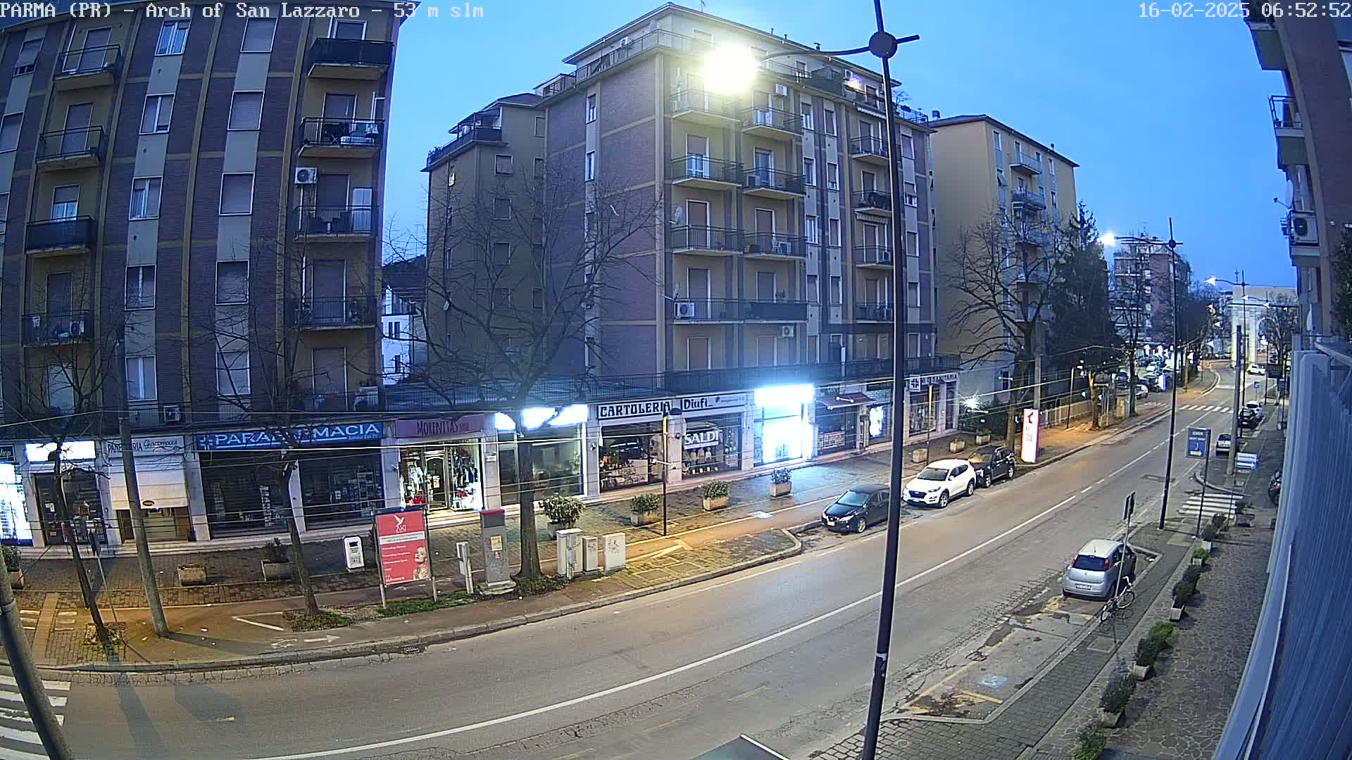 A street scene at twilight shows a multi-story building with shops on the ground floor, cars parked along the side of the road, and a few cars driving down the street.