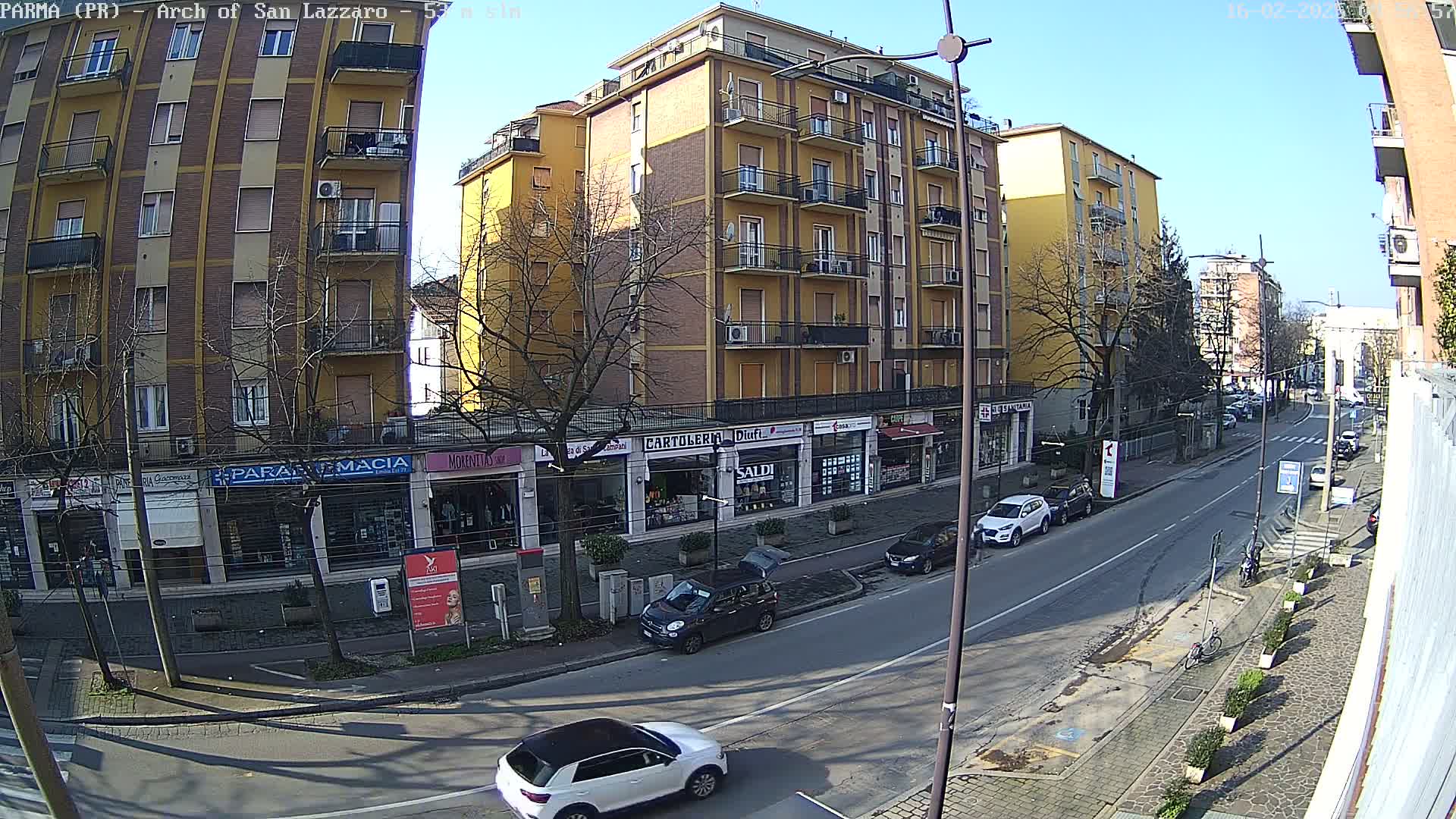 A sunny day on a city street lined with multi-story buildings and shops, with cars parked along the side and a few cars driving down the street.
