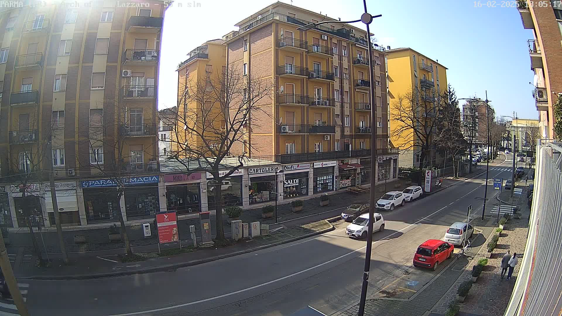 A sunny city street scene shows several multi-story buildings, parked cars, and a few pedestrians.
