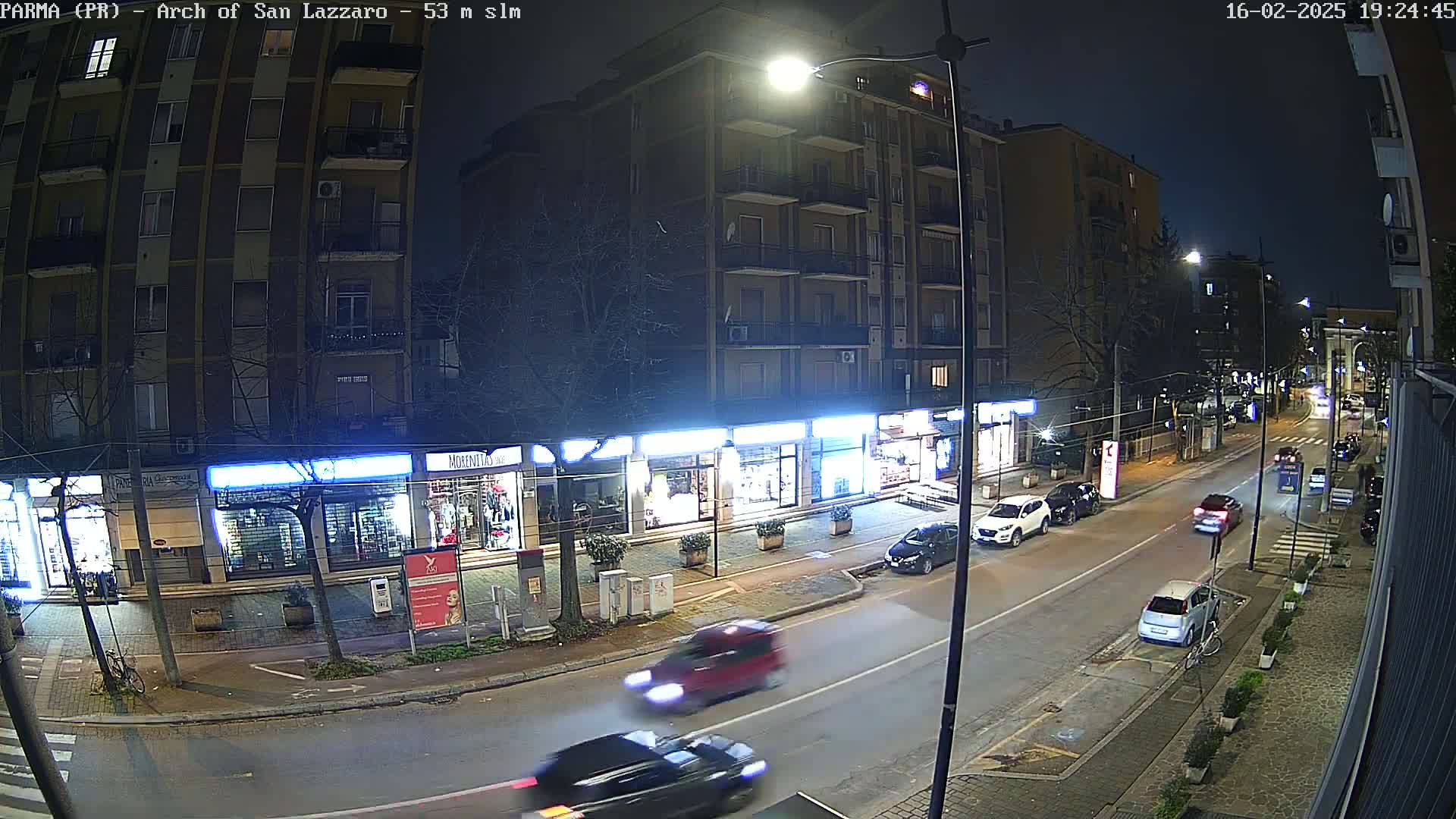 A nighttime street scene shows several cars driving along a road lined with shops and apartment buildings under streetlights.