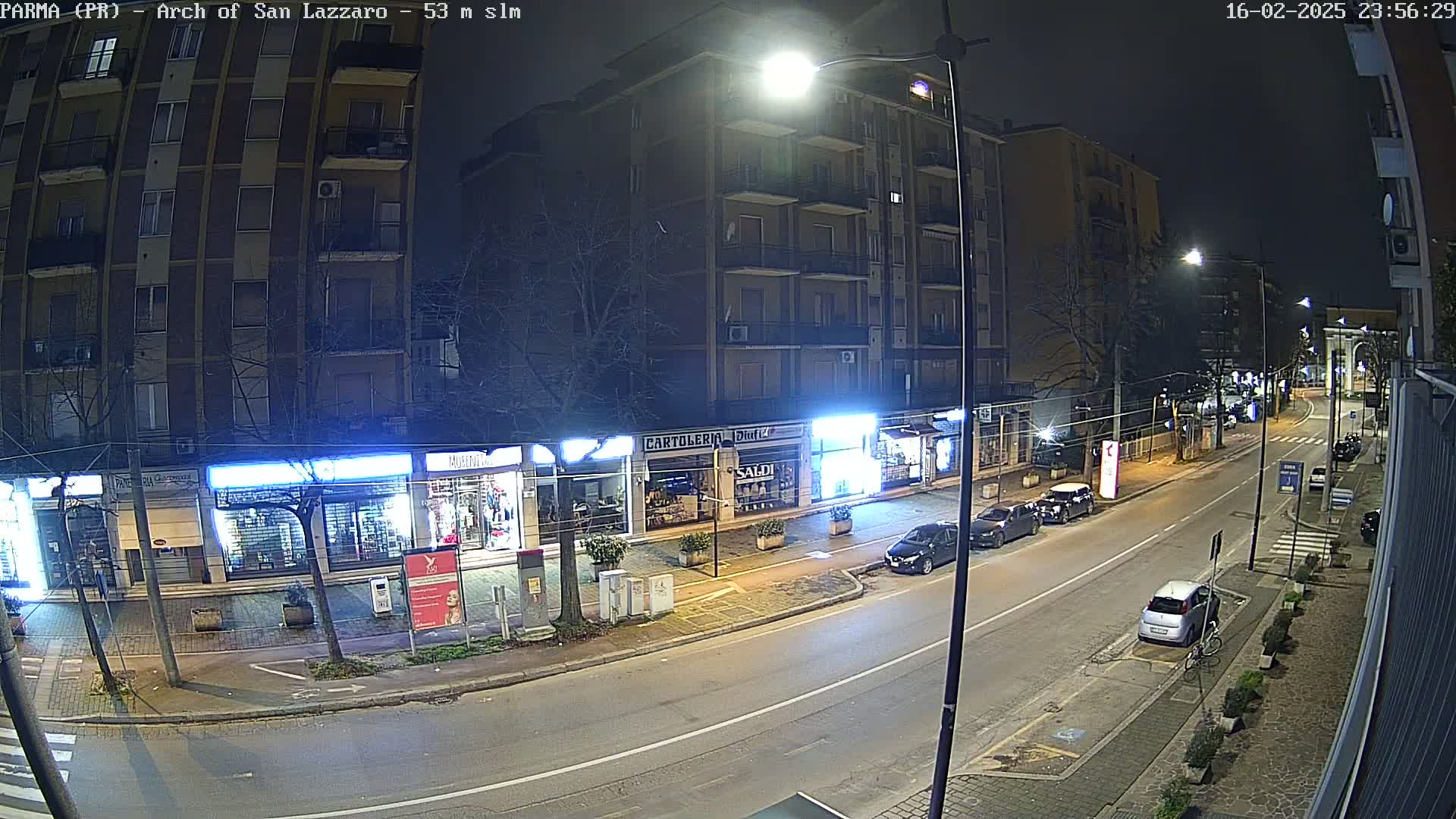 A nighttime street scene shows a mostly empty road in front of several stores and a multi-story building, with a few parked cars and one car driving.