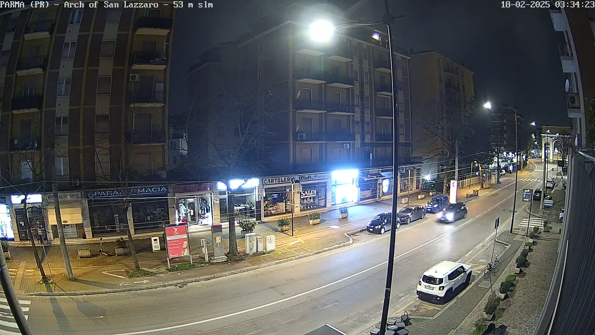 A nighttime street scene shows several cars parked and driving along a road lined with multi-story buildings and shops.