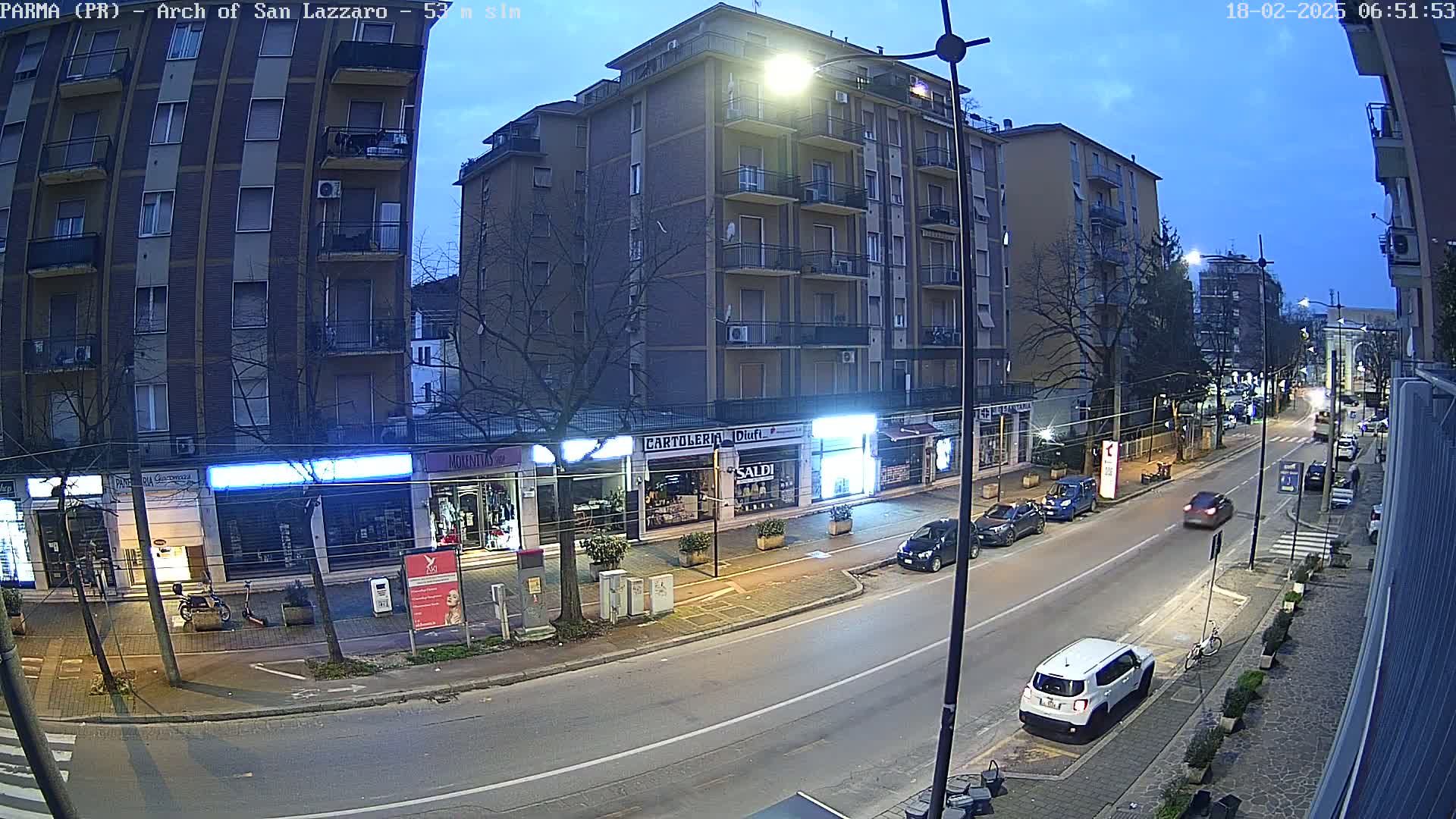 A street scene at twilight shows several parked cars along the curb next to a row of storefronts, with a few cars driving down the street.