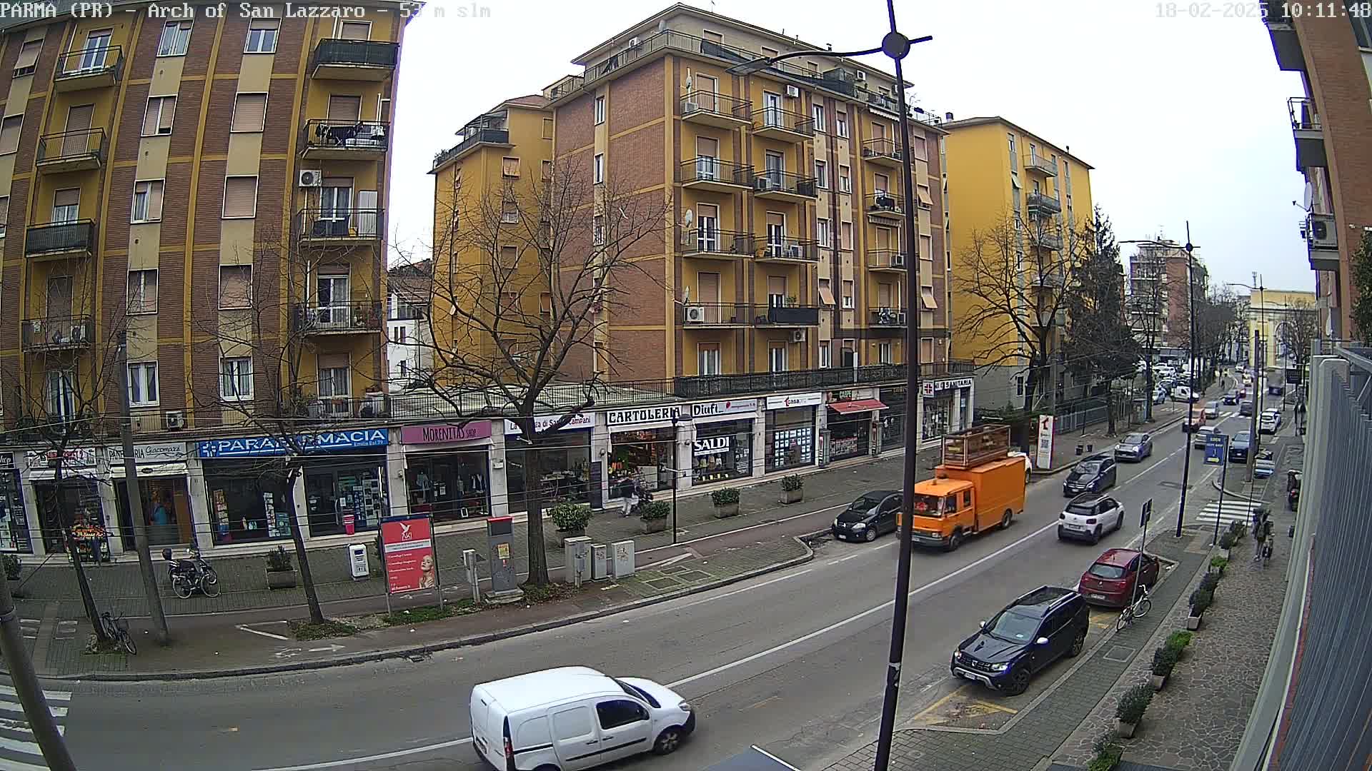 A street scene on an overcast day shows a multi-story yellow building with shops on the ground floor, parked cars, and a few pedestrians, all under a mostly cloudy sky.