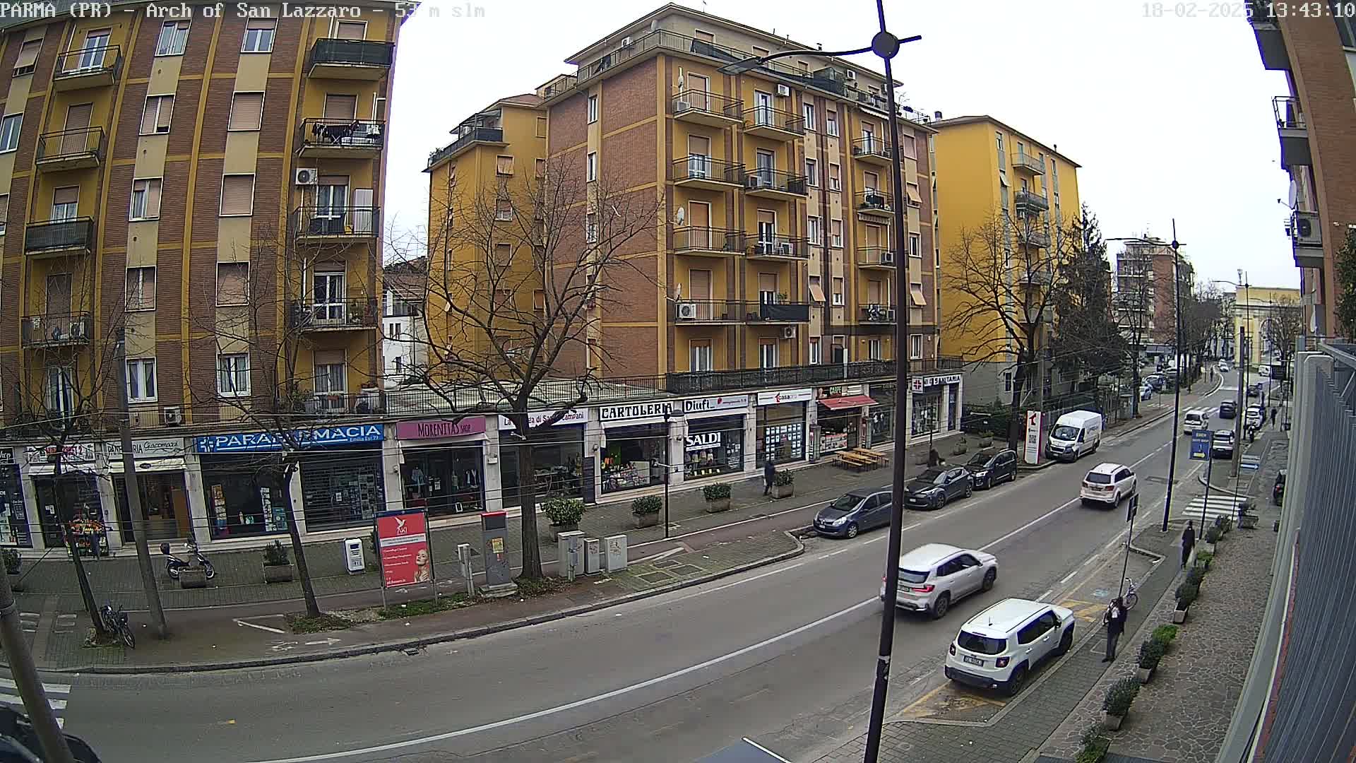 A street scene on an overcast day shows a multi-story yellow building with shops on the ground floor, parked cars, and several pedestrians walking along the sidewalk.