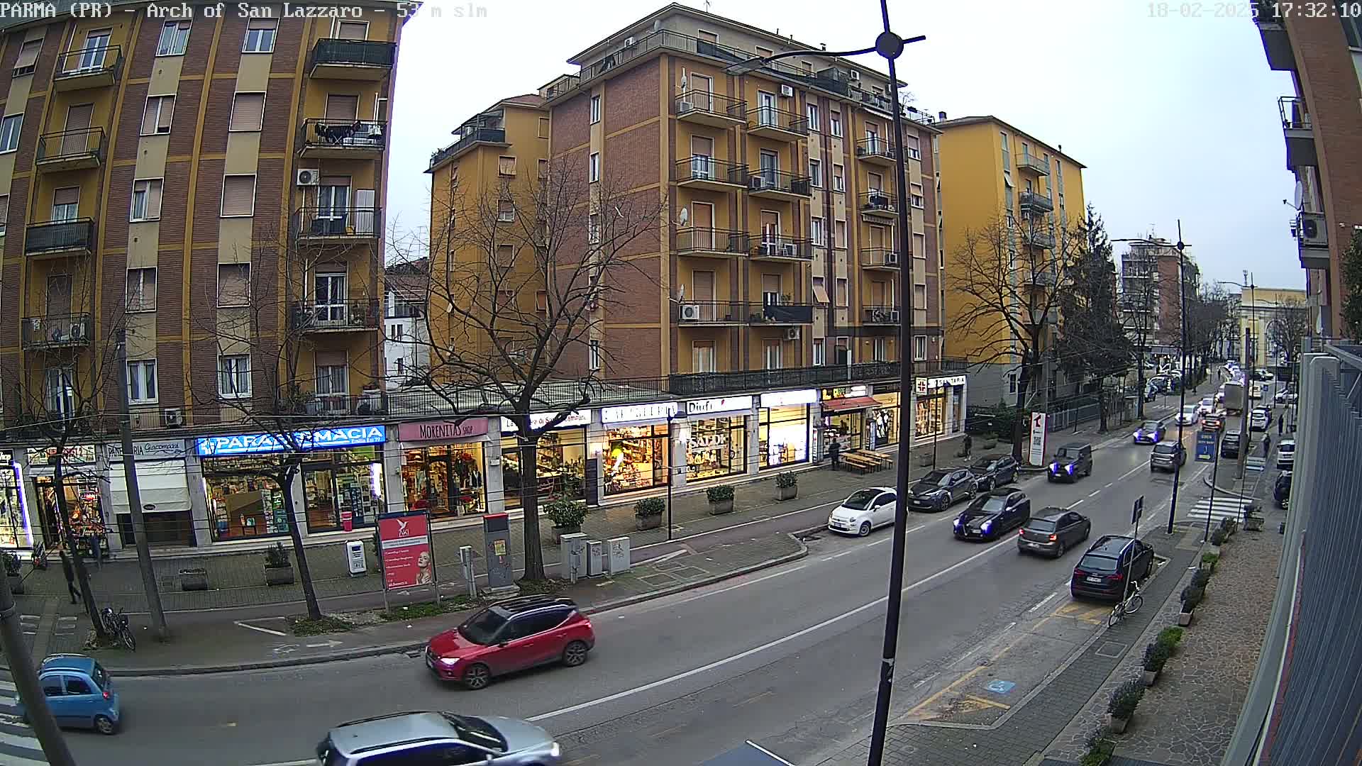 A city street scene on an overcast day shows several cars driving past multi-story buildings with shops on the ground floor.
