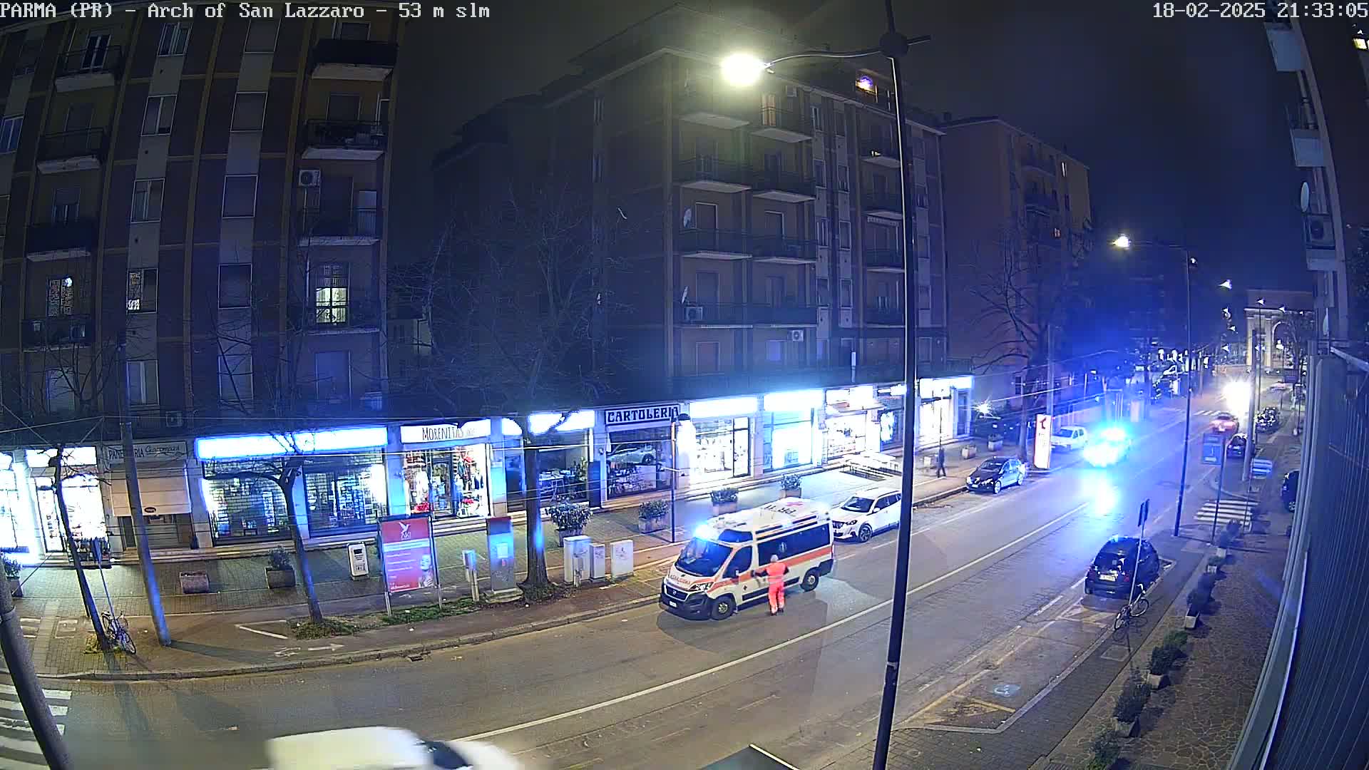 A nighttime street scene shows an ambulance stopped on a city street next to several parked cars, under partly cloudy skies.