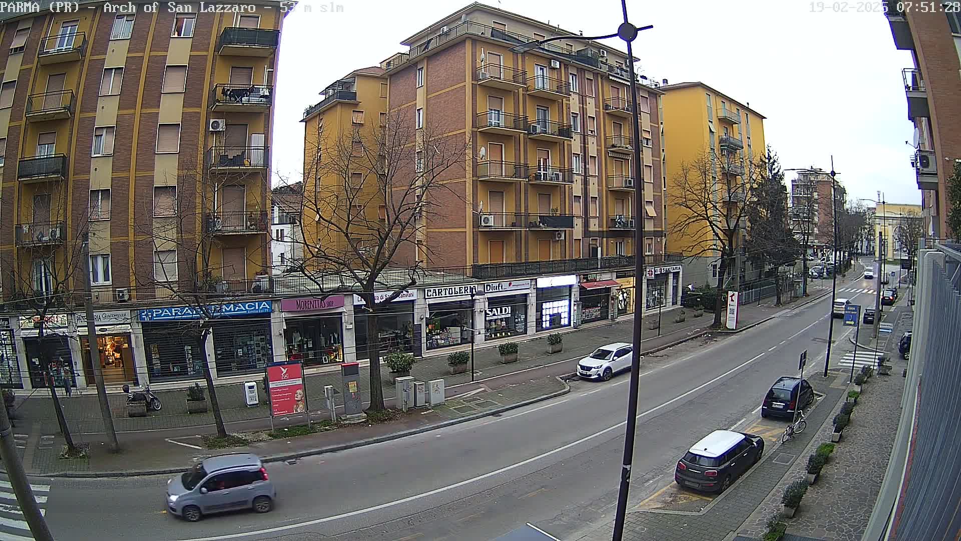 A street scene on an overcast day shows a multi-story yellow building with shops on the ground floor, several parked cars, and sparsely-leafed trees lining the street.