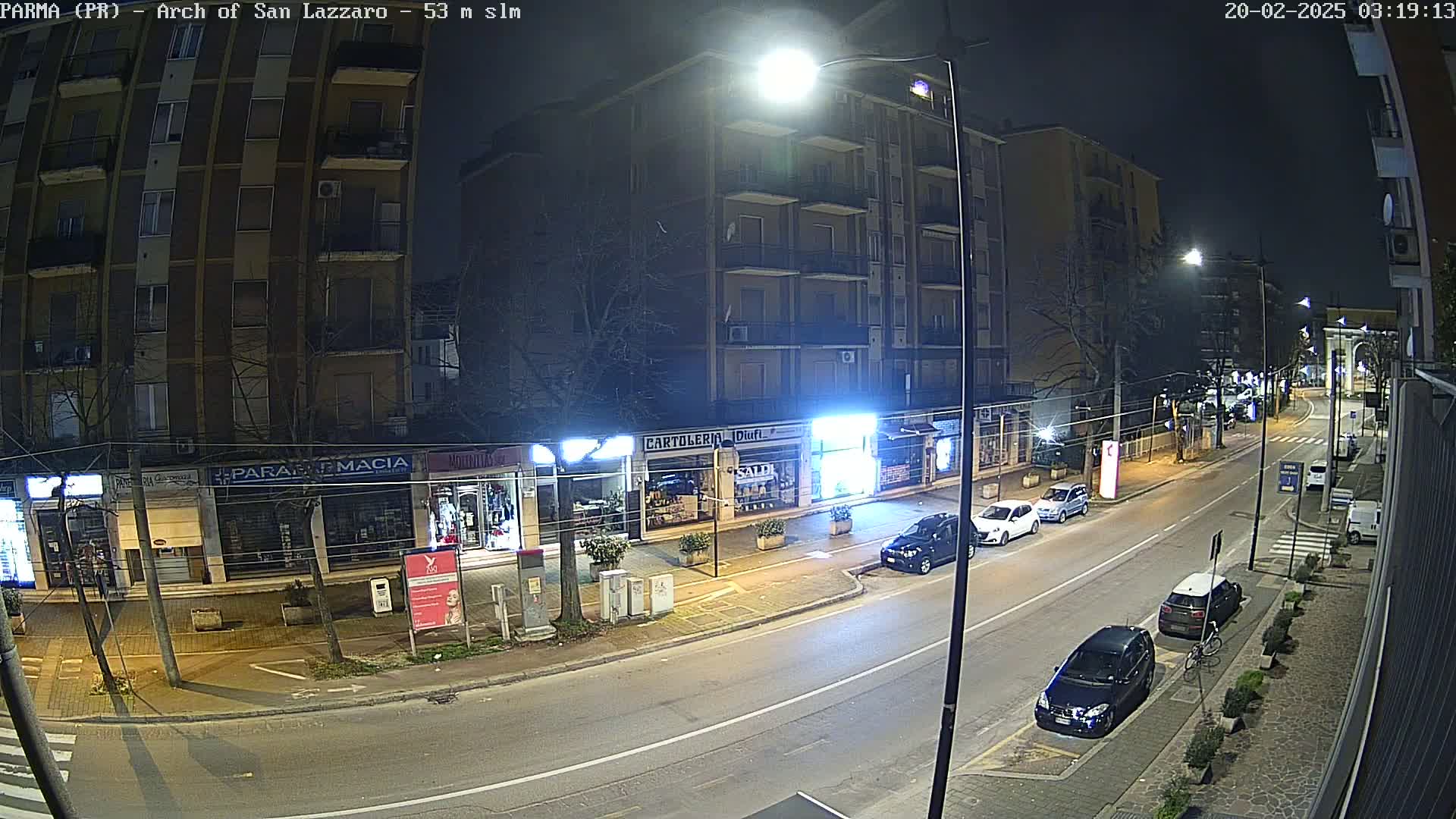 A nighttime street scene shows several parked cars along a road lined with storefronts and apartment buildings under bright streetlights.