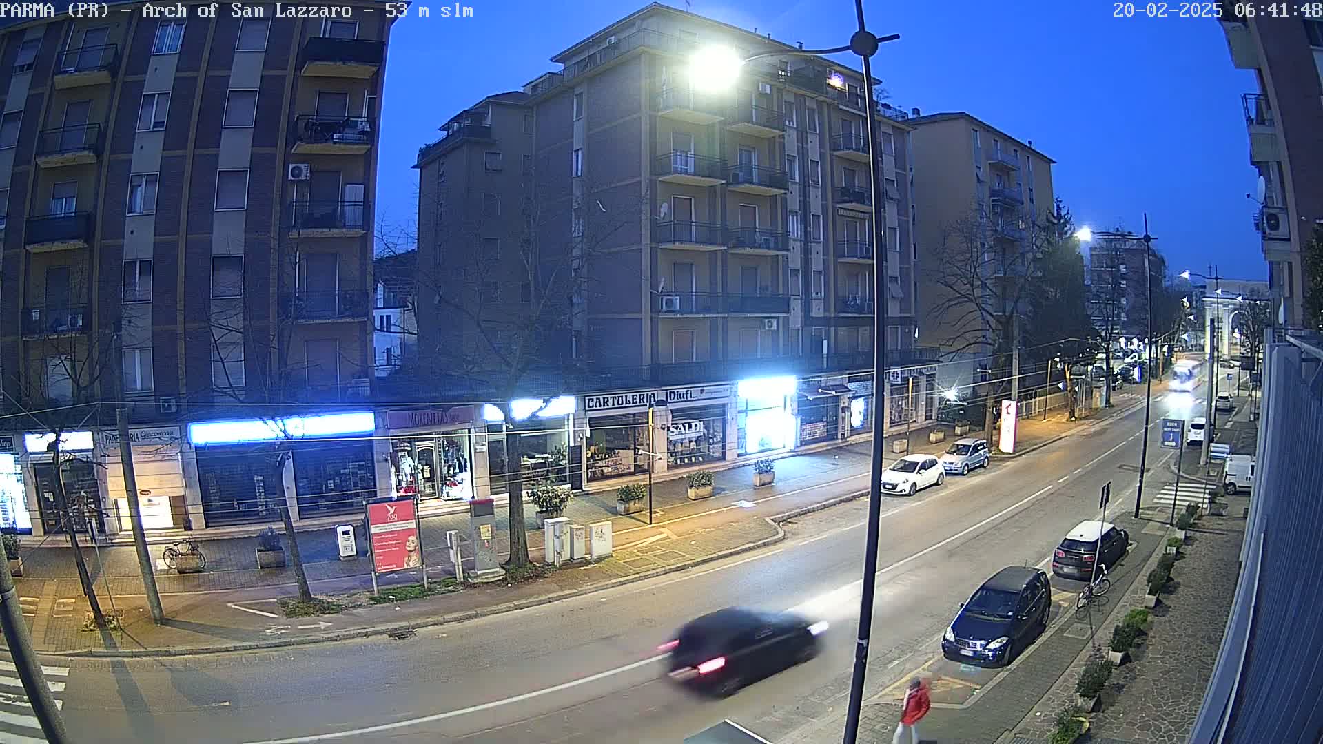 A nighttime street scene shows a multi-story building with shops on the ground floor, several cars parked and driving on the road, and bare trees lining the sidewalk under a twilight sky.
