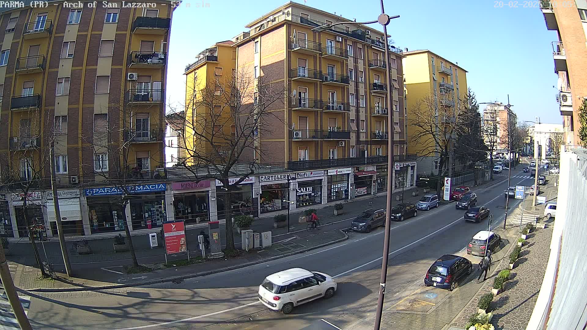 A sunny city street scene shows a row of multi-story buildings with shops on the ground floor, and several cars parked and driving along the street.