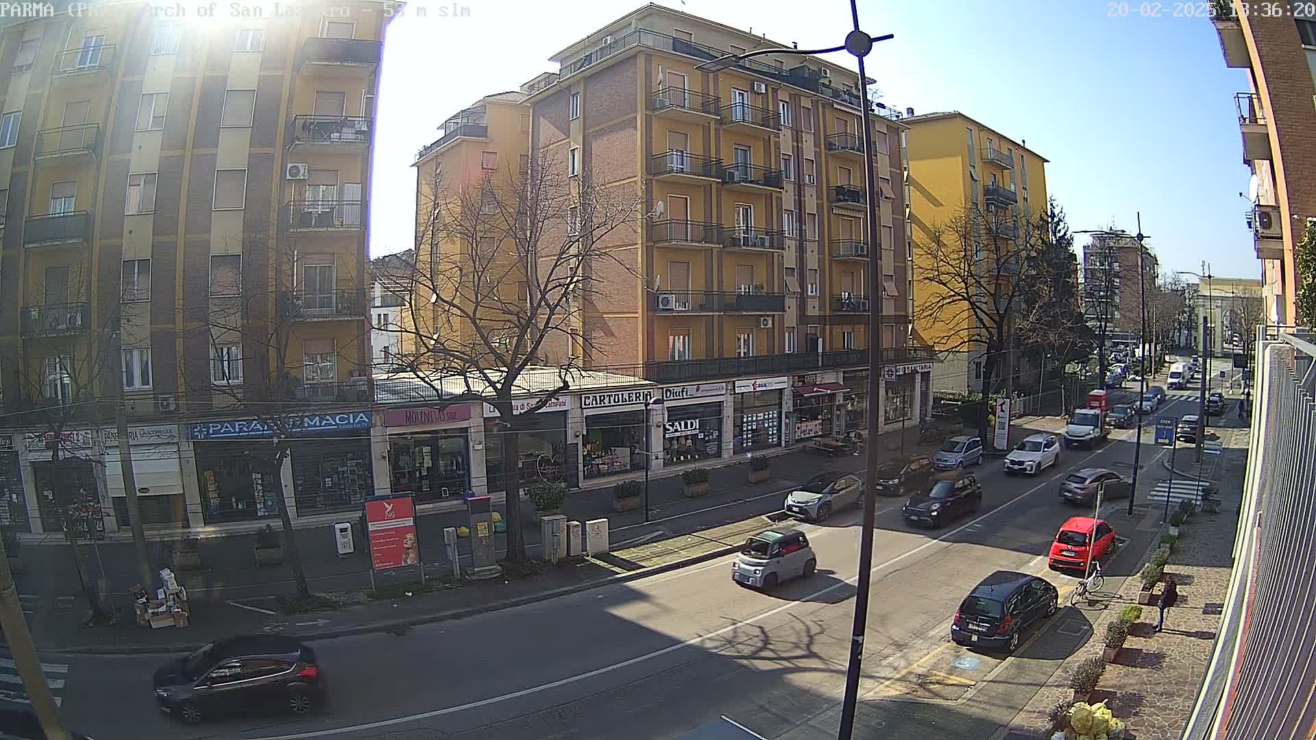 A sunny street scene shows a multi-story yellow building with shops on the ground floor, several cars parked along the street and more cars driving down the road.