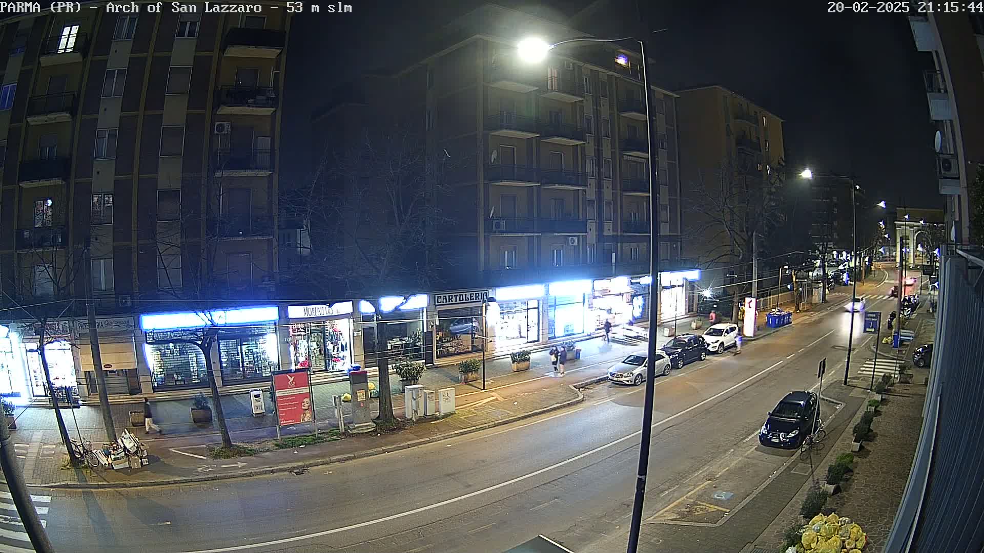 A nighttime street scene shows several parked cars and a few driving cars along a city street lined with storefronts.