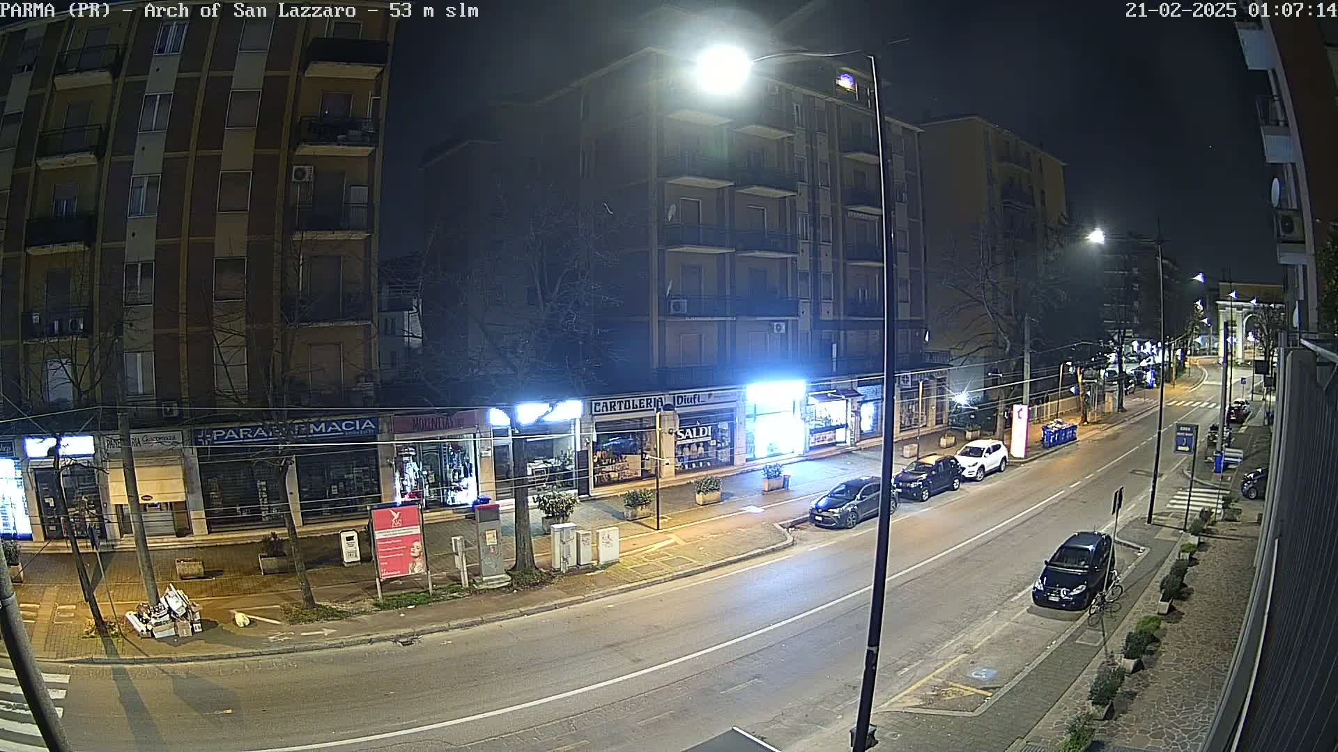 A nighttime street scene shows several parked cars along a road lined with shops and apartment buildings under bright streetlights.