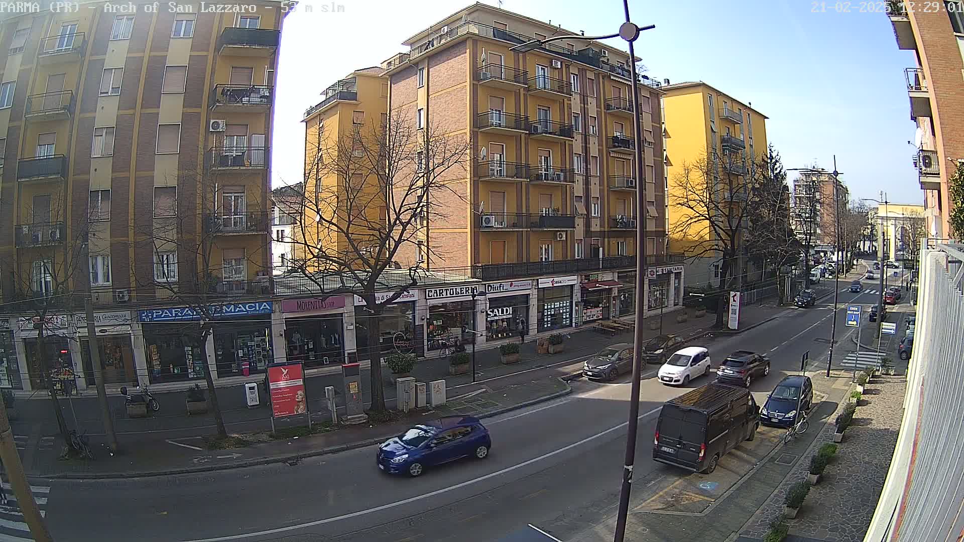 A sunny day view of a city street with several cars parked and driving, alongside multi-story buildings containing shops and a pharmacy.