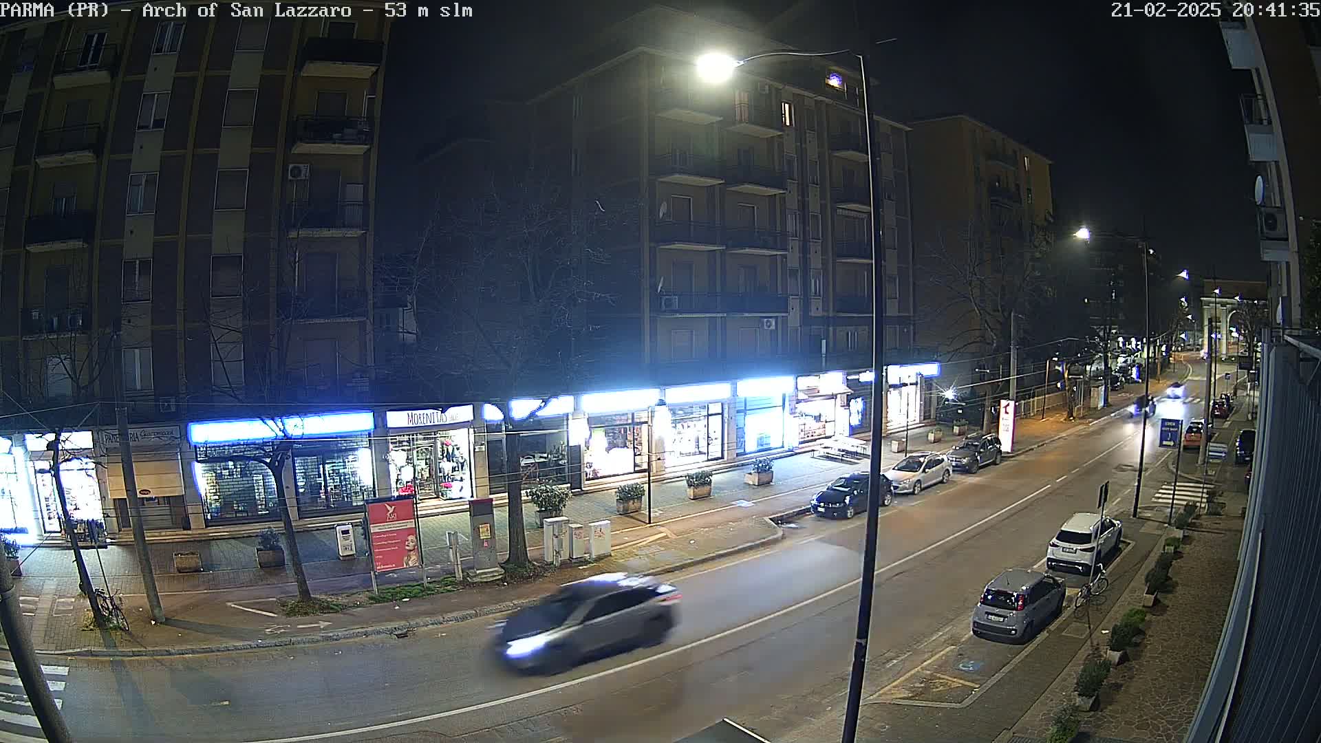 A nighttime street scene shows several parked and moving cars in front of illuminated storefronts and a multi-story building.