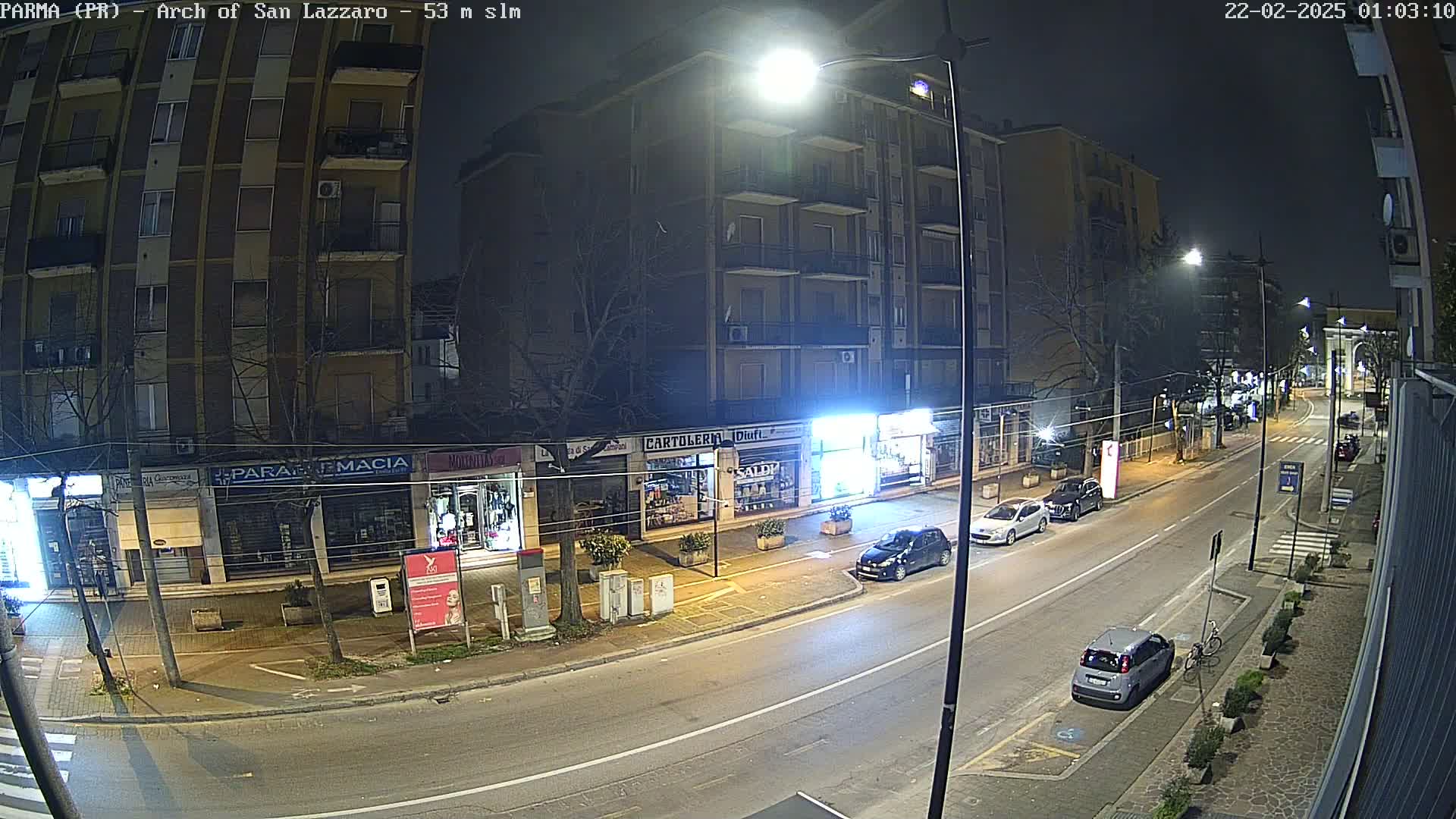 A nighttime street scene shows a row of shops and apartment buildings under bright streetlights, with a few parked cars and a car driving down the road.