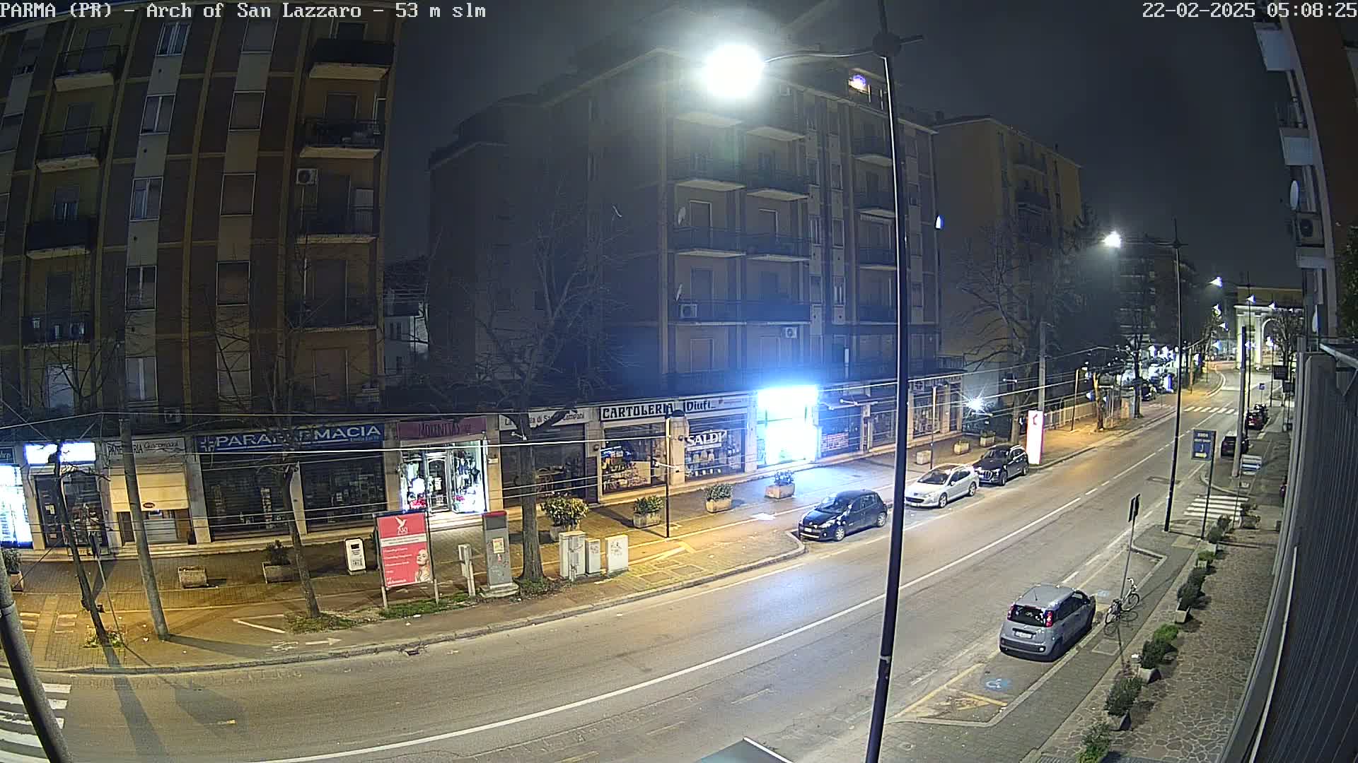 A nighttime street scene shows several cars parked along a roadway in front of shops and apartment buildings under bright streetlights.