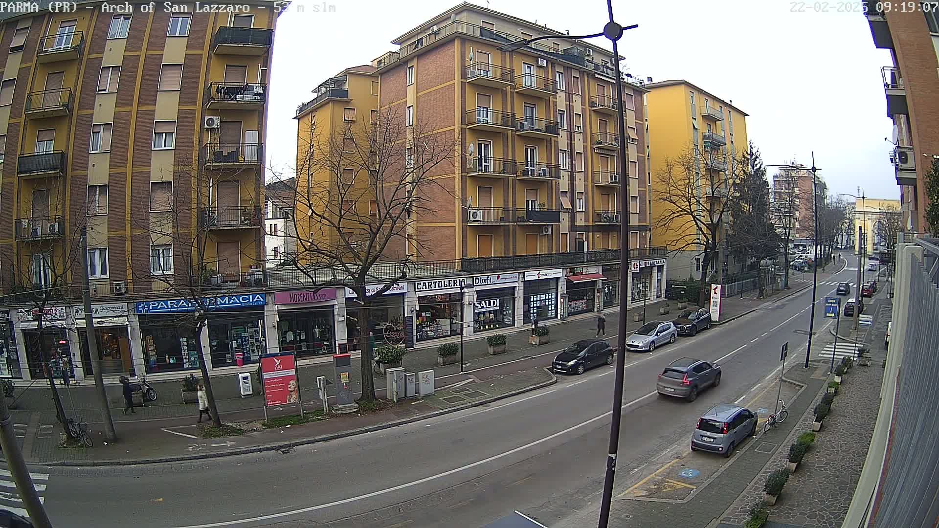 A street scene on a cloudy day shows a multi-story yellow building with shops on the ground floor, several parked cars, and pedestrians on a sidewalk.