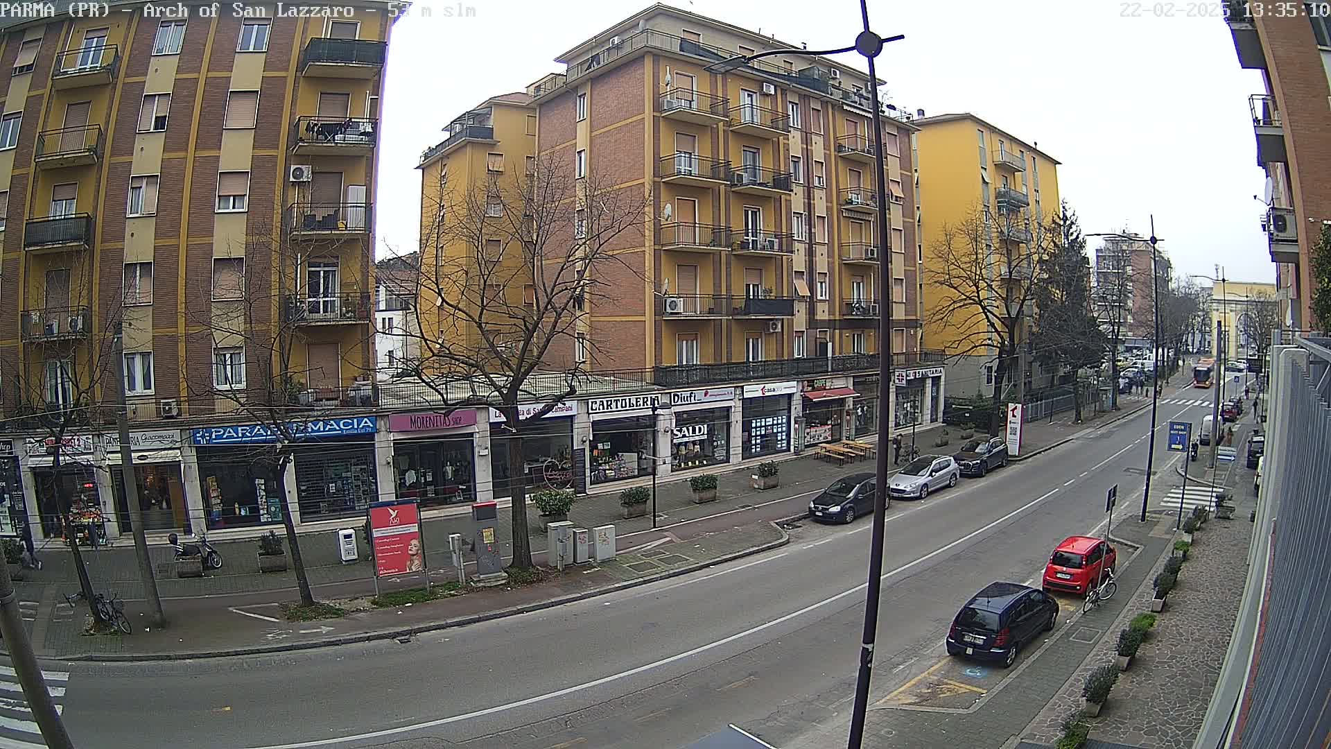 A street scene on an overcast day shows a multi-story yellow building with shops on the ground floor, several parked cars, and a few people walking along the sidewalk.