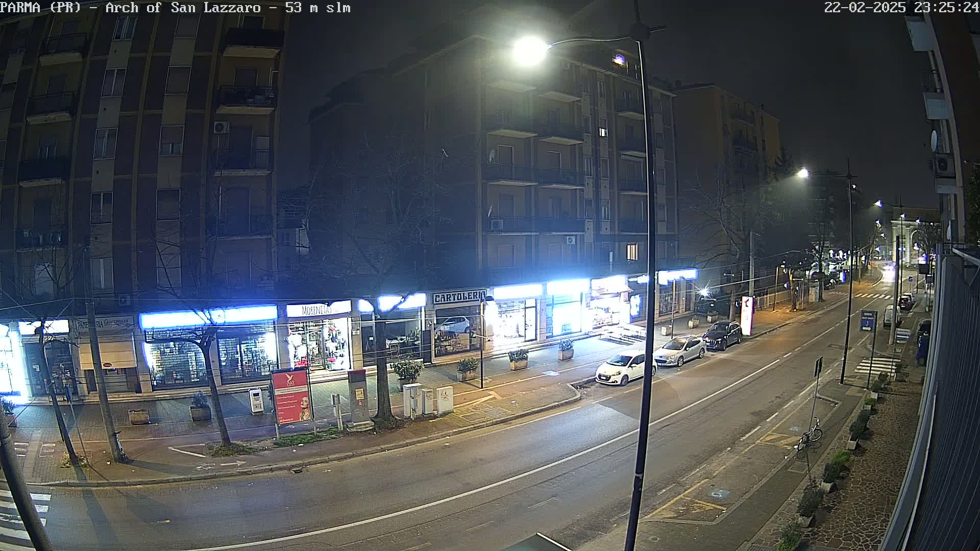 A nighttime street scene shows several parked cars in front of shops under bright streetlights, with a few cars driving down the road on a slightly hazy night.