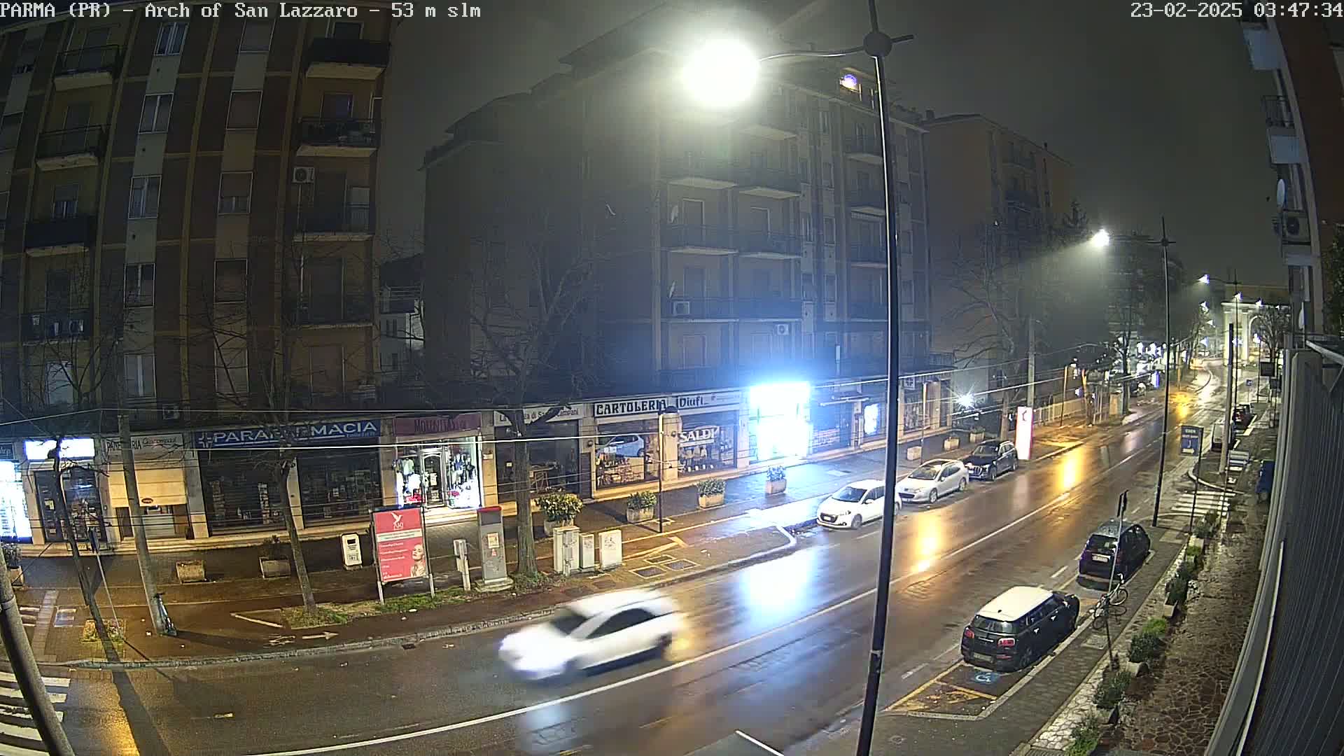 A wet, nighttime street scene shows several cars driving past buildings with storefronts under bright streetlights.